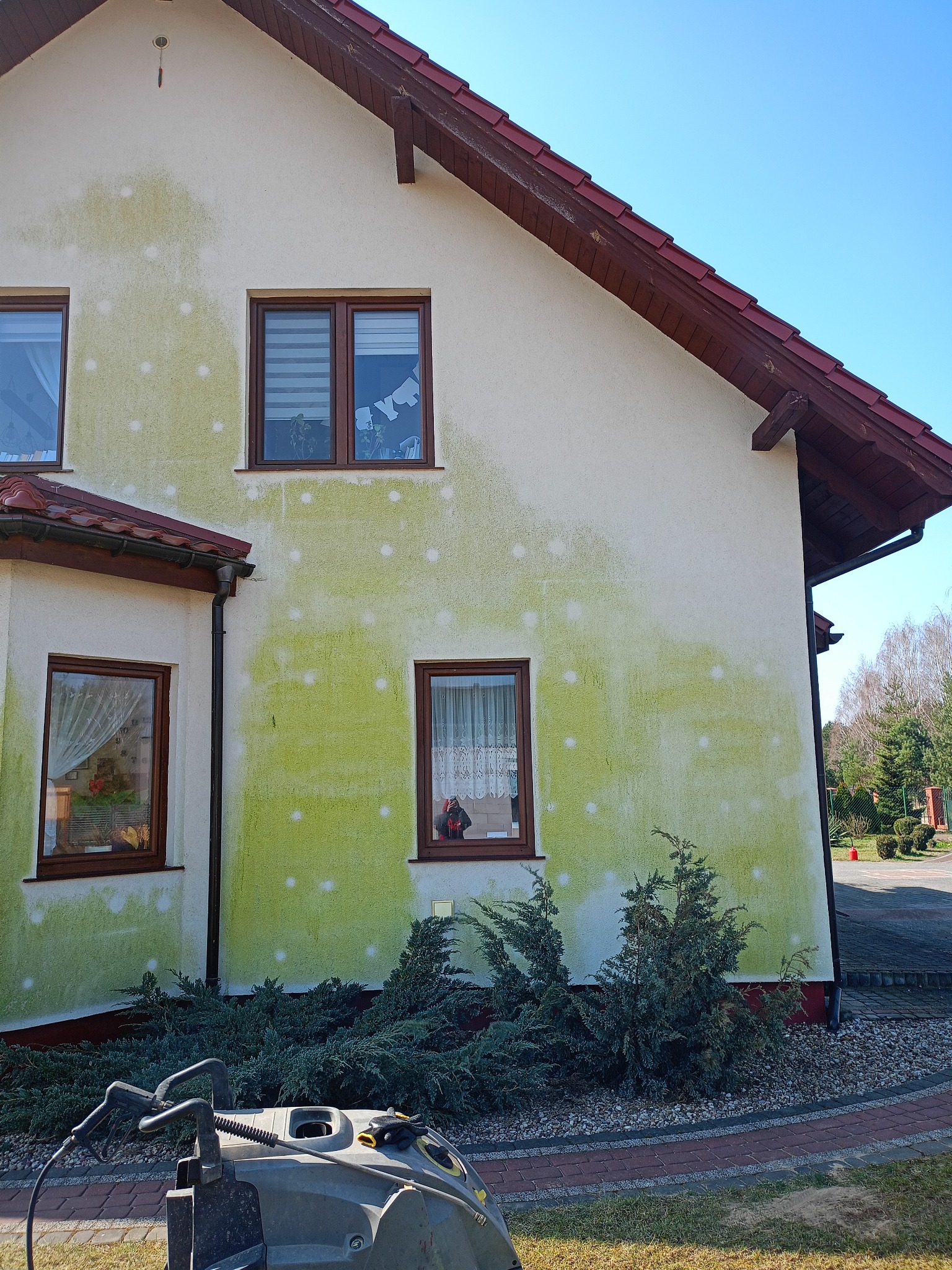 Elewacja domu z widocznymi zielonymi zabrudzeniami i plamami po czyszczeniu, z myjką ciśnieniową na pierwszym planie.