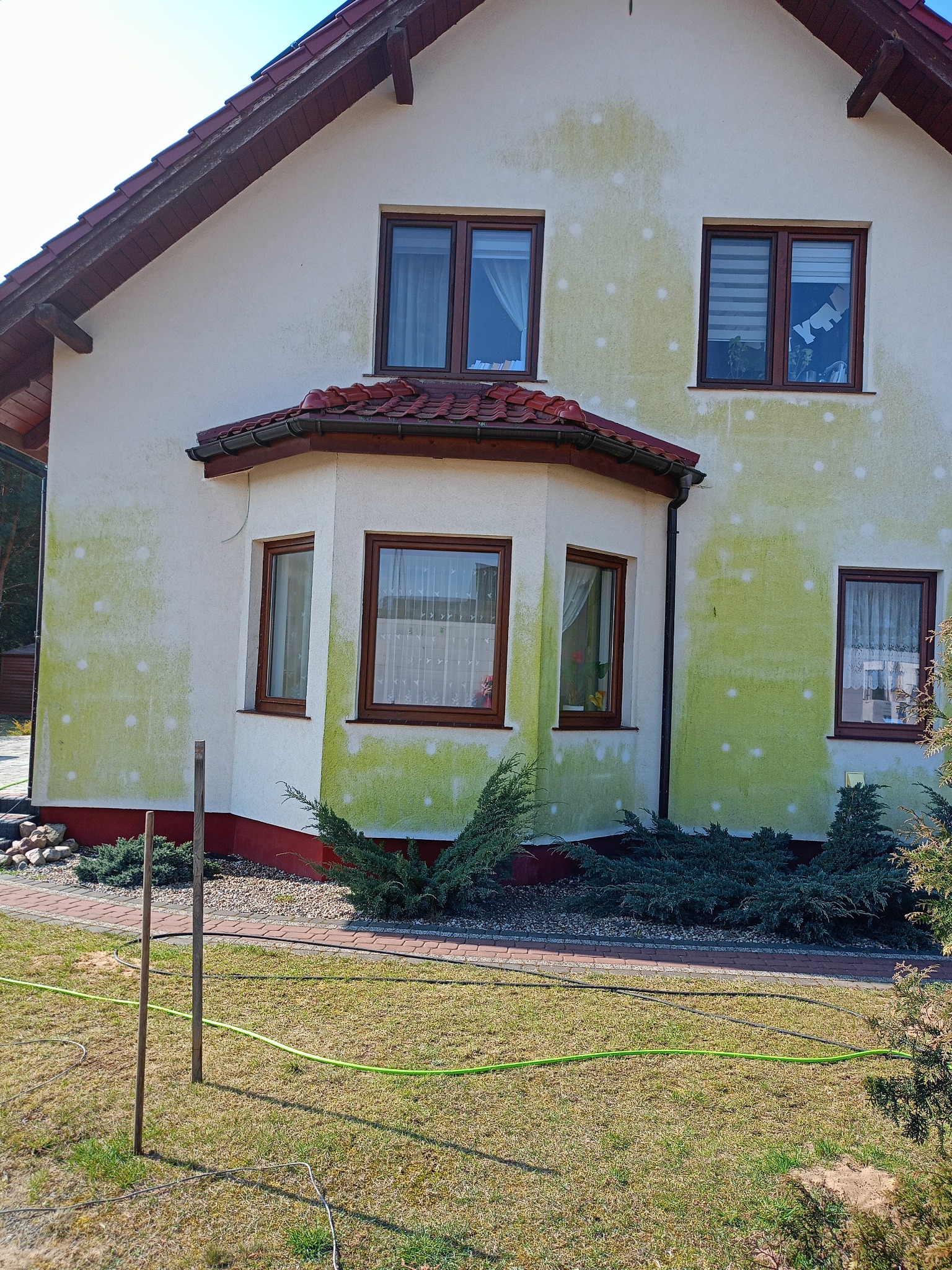 Elewacja domu z widocznymi plamami i punktami, przygotowana do malowania, z widocznym trawnikiem i elementami ogrodu.