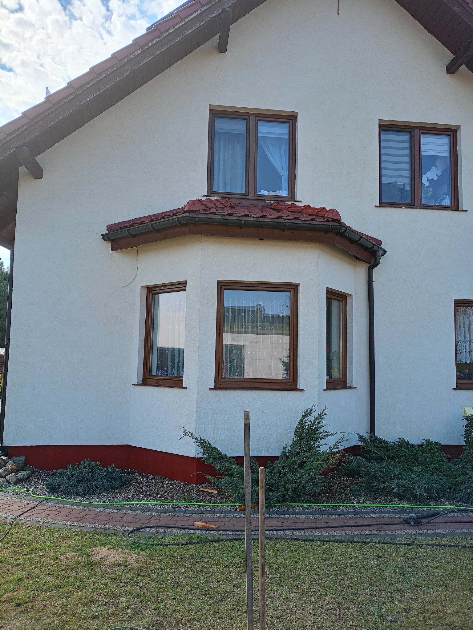 Dom jednorodzinny z białymi ścianami zewnętrznymi i brązowymi oknami, widoczne elementy elewacji po renowacji, w tle błękitne niebo z chmurami.
