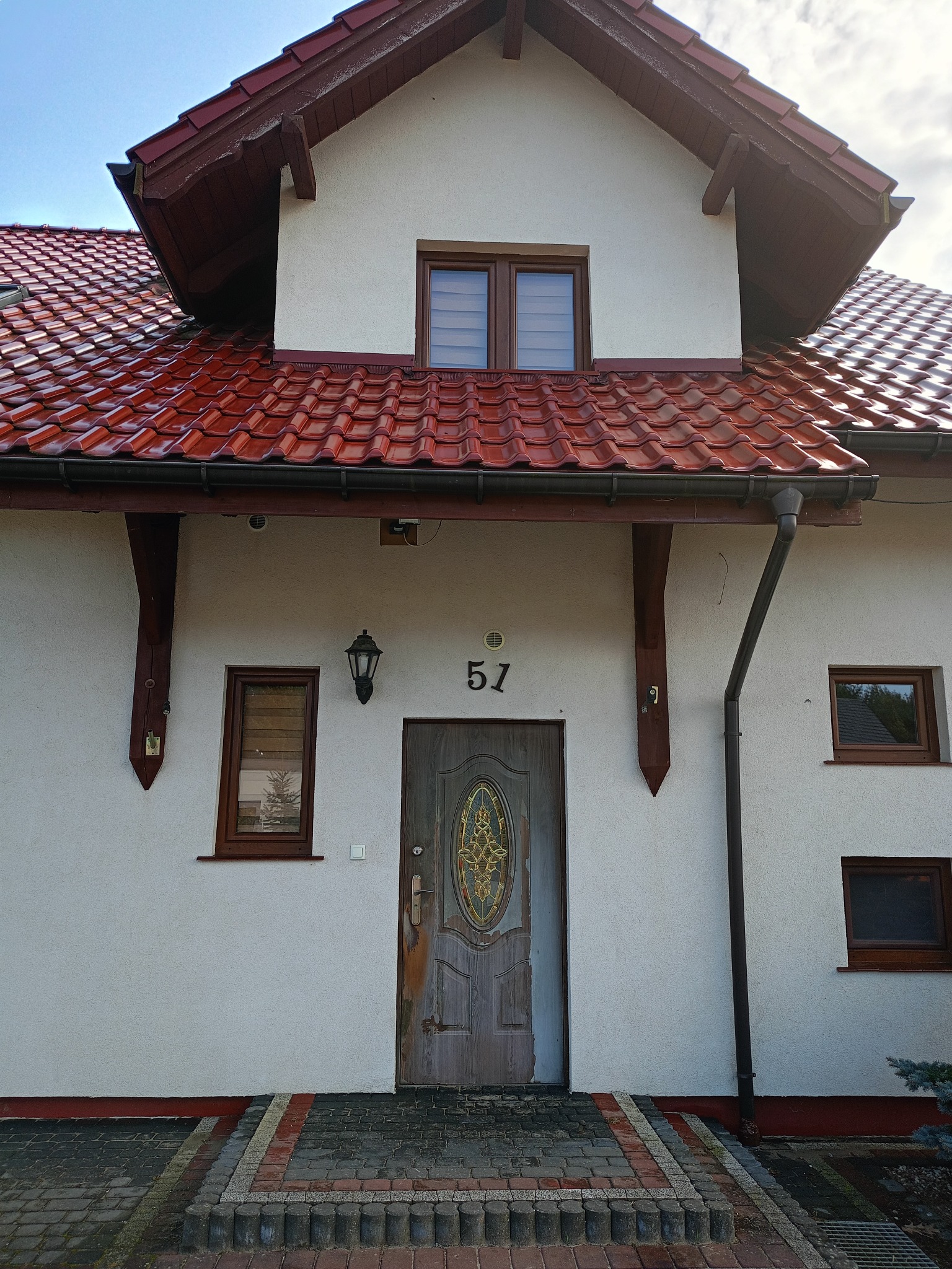 Front domu jednorodzinnego z czerwoną dachówką, numerem 51 nad drewnianymi drzwiami z witrażem, widoczne okna z brązowymi ramami i fragment elewacji.