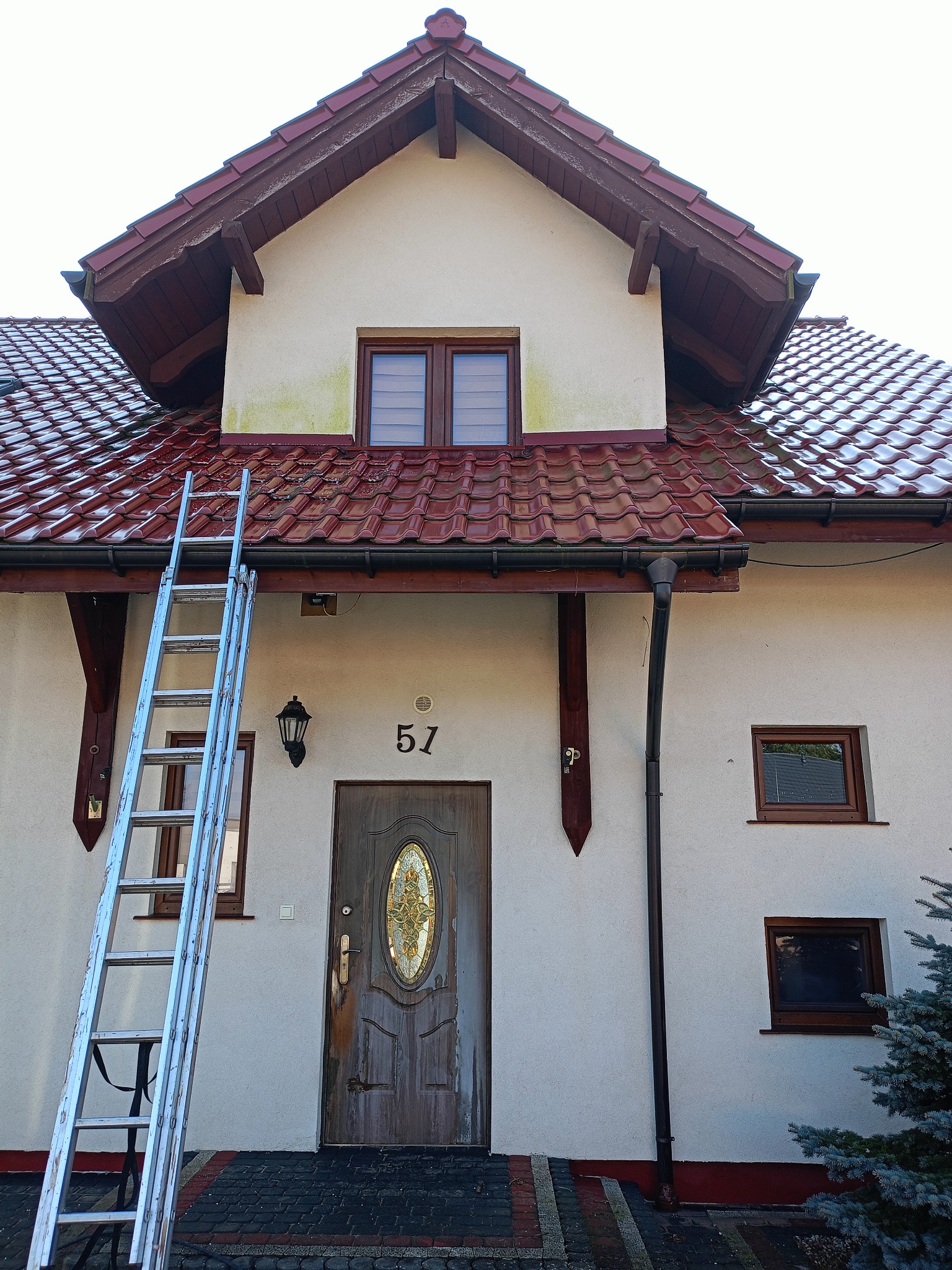 Dwupiętrowy dom z czerwonym dachem, drabina oparta o elewację, widoczne ślady zabrudzeń i wilgoci na ścianie pod oknem na poddaszu.