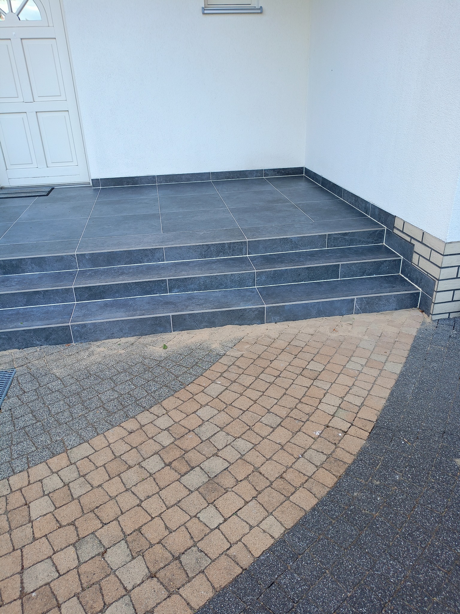 Wykończone płytkami schody wejściowe do budynku, z widocznym fragmentem chodnika z kostki brukowej. Detal wykończenia elewacji przy schodach.