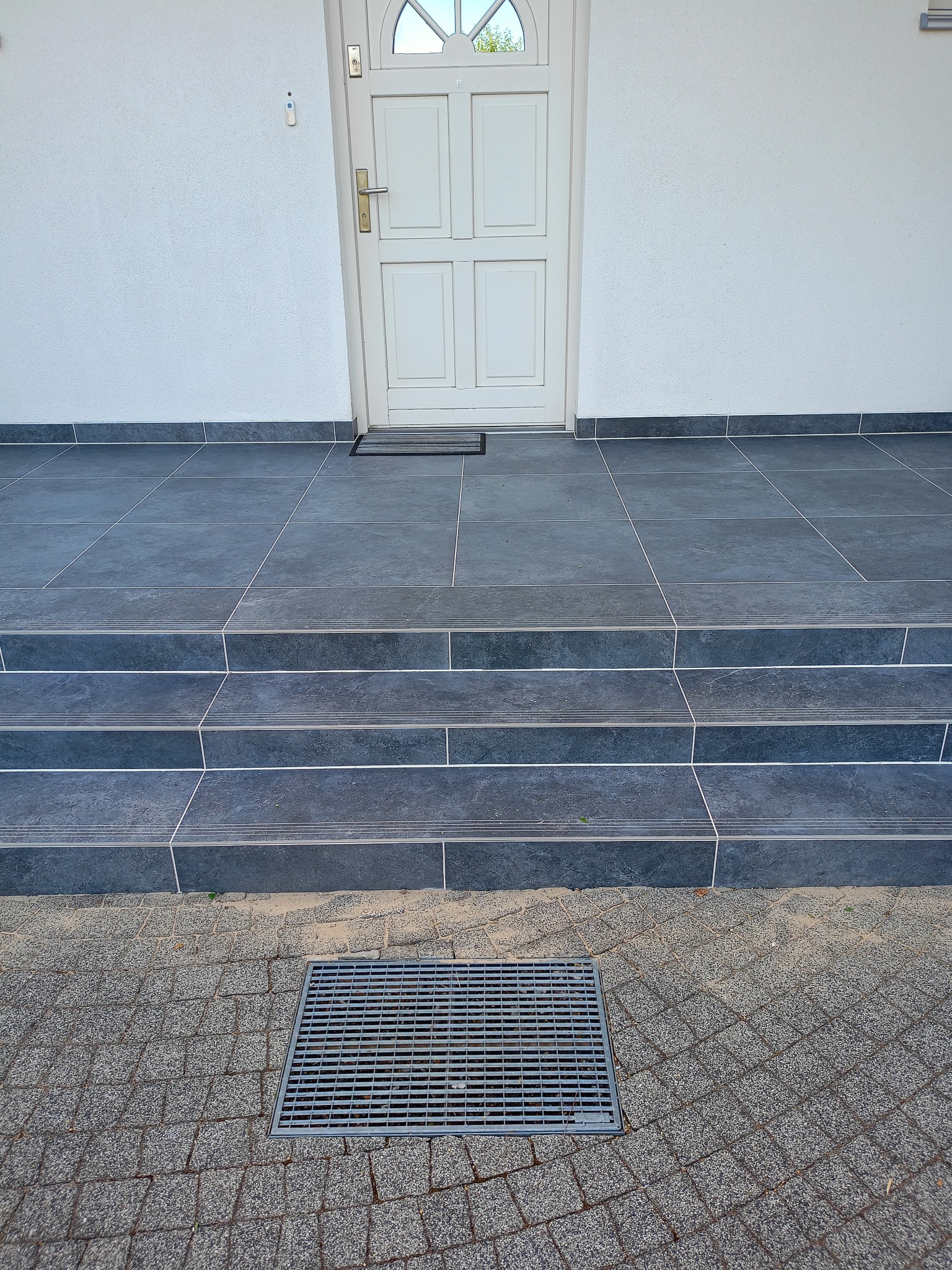 Wykończone płytkami schody wejściowe do domu z białymi drzwiami i kratką odpływową. Ciemnoszare płytki z jasnymi fugami. Widok na elewację i chodnik z kostki.