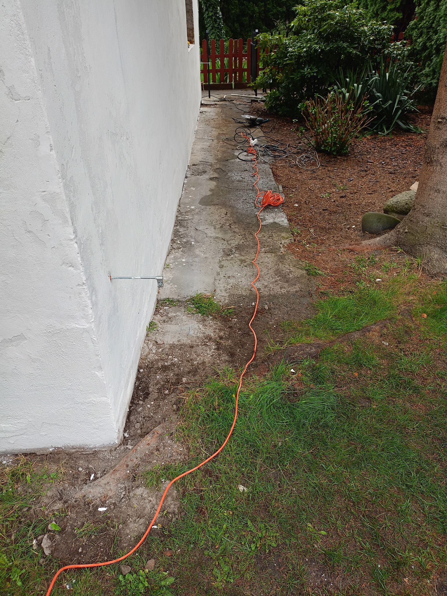 Biała ściana budynku z zamontowanym wspornikiem. Na ziemi leży pomarańczowy kabel zasilający i narzędzia, co sugeruje prace elewacyjne w toku.