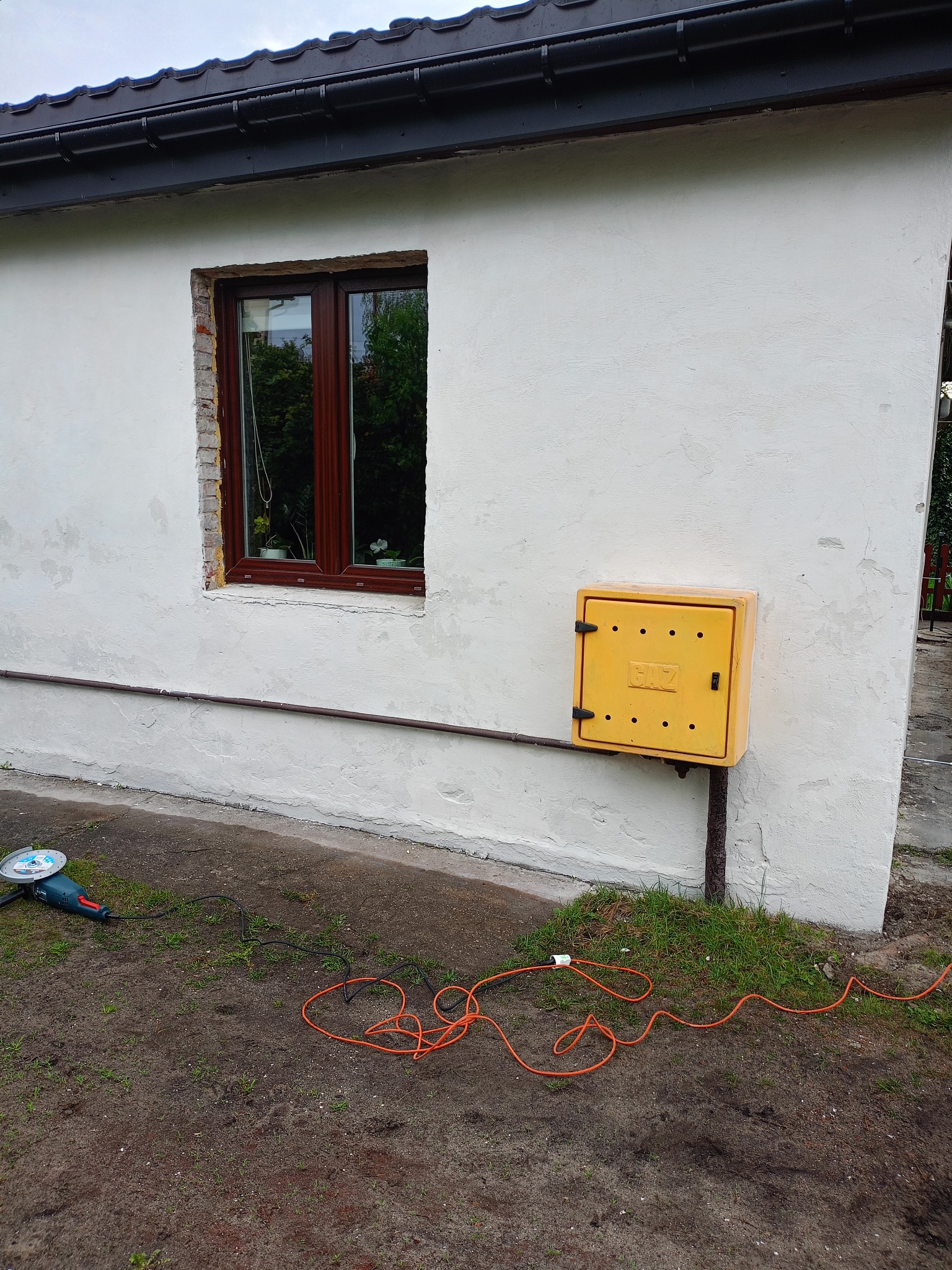 Fragment elewacji z oknem w trakcie wymiany i żółtą skrzynką gazową. Na ziemi leży szlifierka kątowa i pomarańczowy kabel.