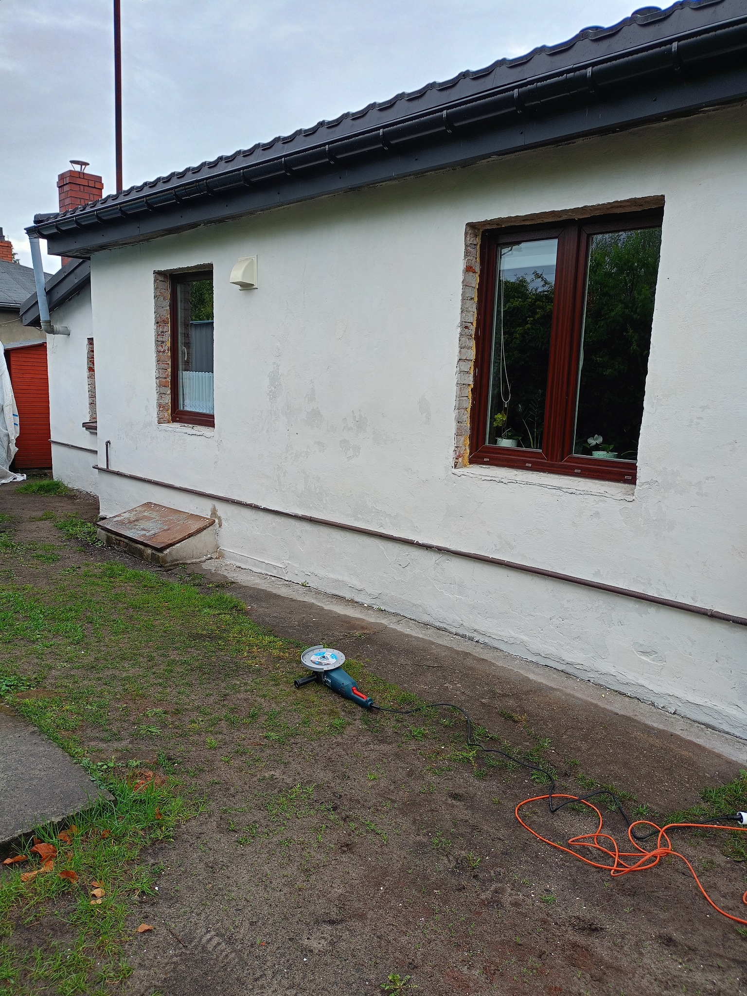 Biała elewacja domu z oknami w brązowych ramach, widoczne ślady po kuciu, szlifierka kątowa na trawie, kabel elektryczny. Przygotowania do prac elewacyjnych.