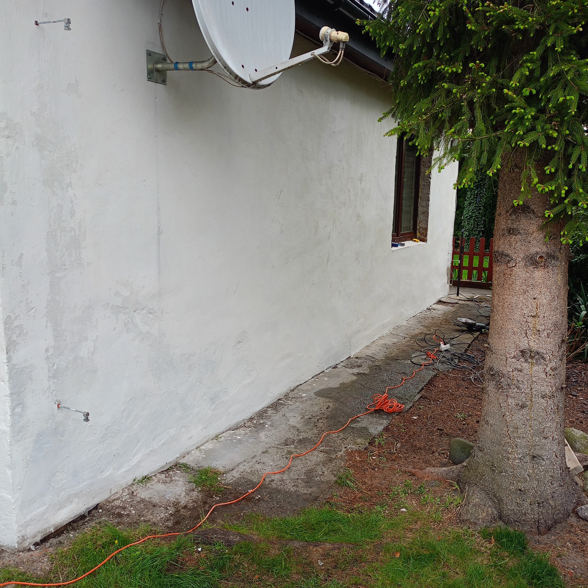 Biała elewacja budynku z anteną satelitarną, widoczne narzędzia i przewody, obok rośnie świerk. Świeżo nałożony tynk, nierówna faktura.