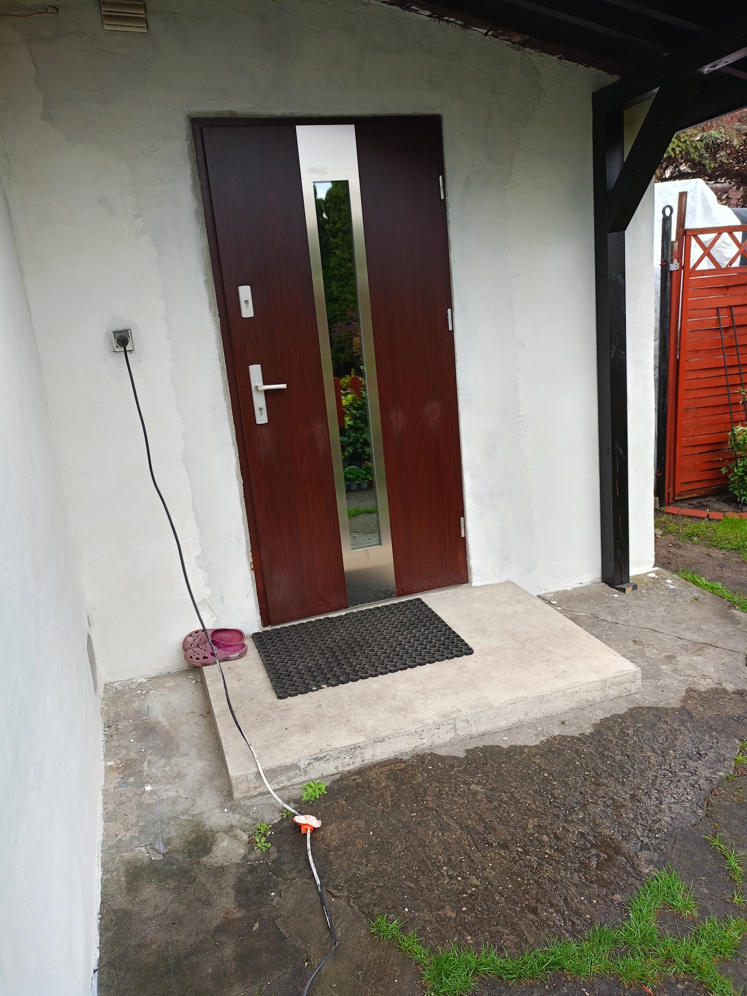 Wejście do domu z brązowymi drzwiami z metalową wstawką, widoczne ślady świeżego tynku na ścianie, przed wejściem leżą klapki i kabel. Prace wykończeniowe.