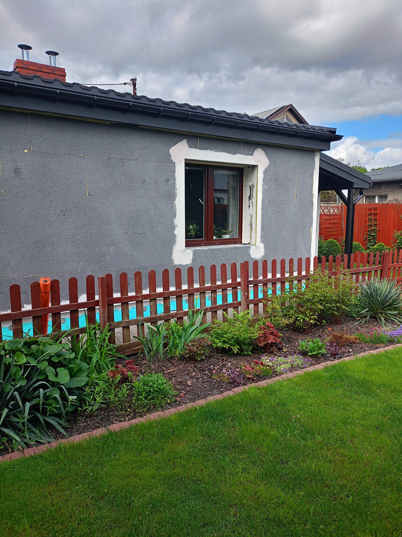 Szary dom w trakcie ocieplania, widoczna izolacja pod oknem i rolka pomarańczowej siatki. Czerwone ogrodzenie i zadbany ogród z przodu.