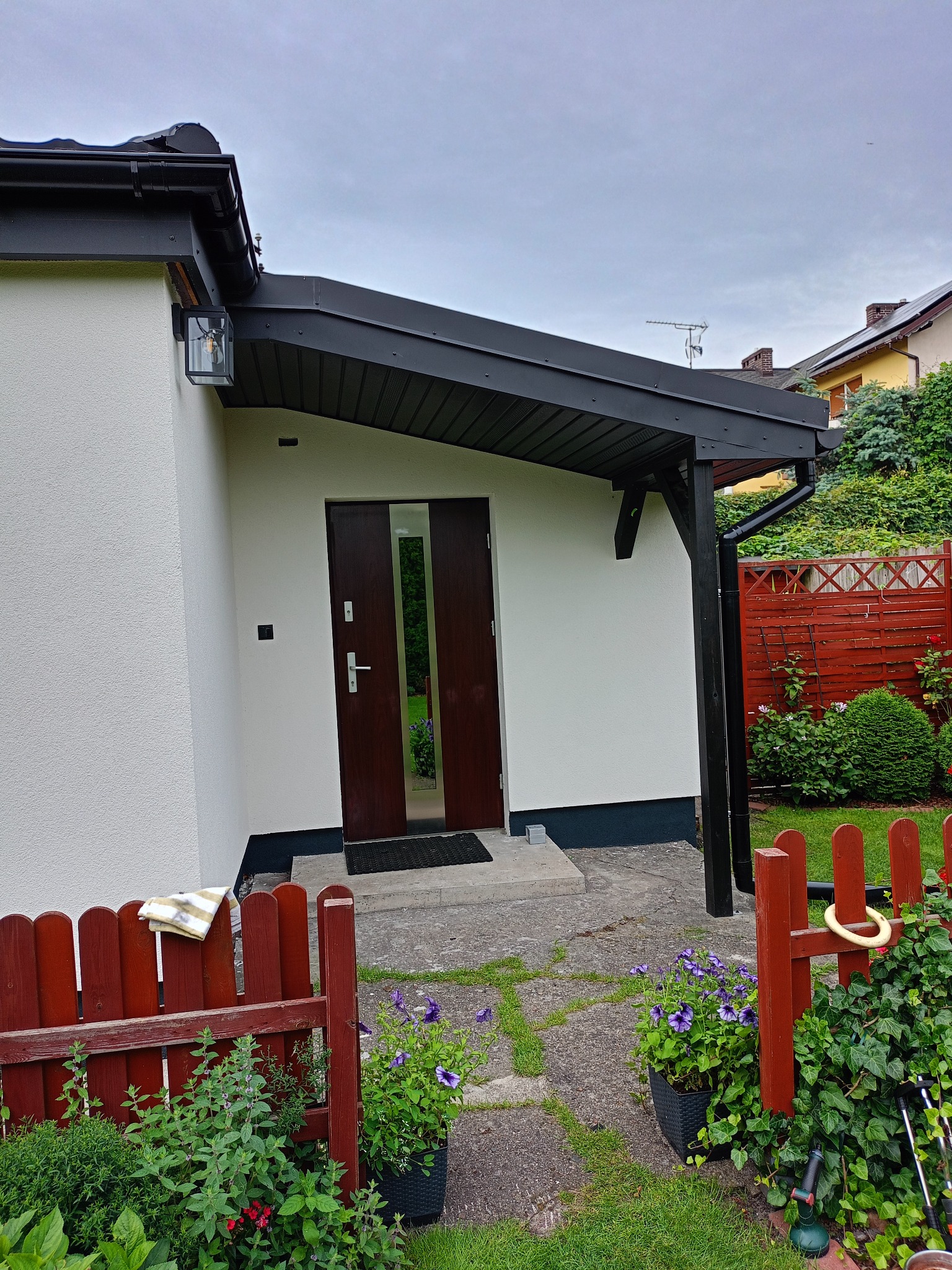 Biała elewacja domu z czarnym zadaszeniem nad wejściem i brązowymi drzwiami. Widoczne rośliny w donicach i fragment czerwonego płotu. Oświetlenie zewnętrzne.