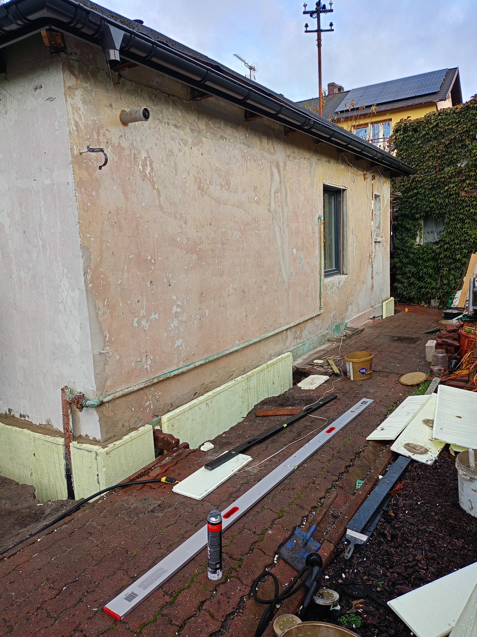 Przygotowania do ocieplenia elewacji budynku z widocznymi płytami izolacyjnymi, narzędziami i fragmentem elewacji przed nałożeniem izolacji. Widok z poziomu gruntu.