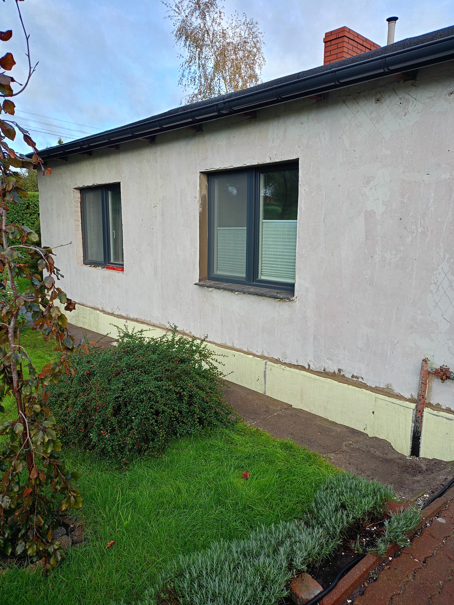 Niski budynek z oknami przed ociepleniem elewacji, widoczne ślady po siatce i żółty styropian przy fundamencie. Fragment ogrodu z zielenią w kadrze.