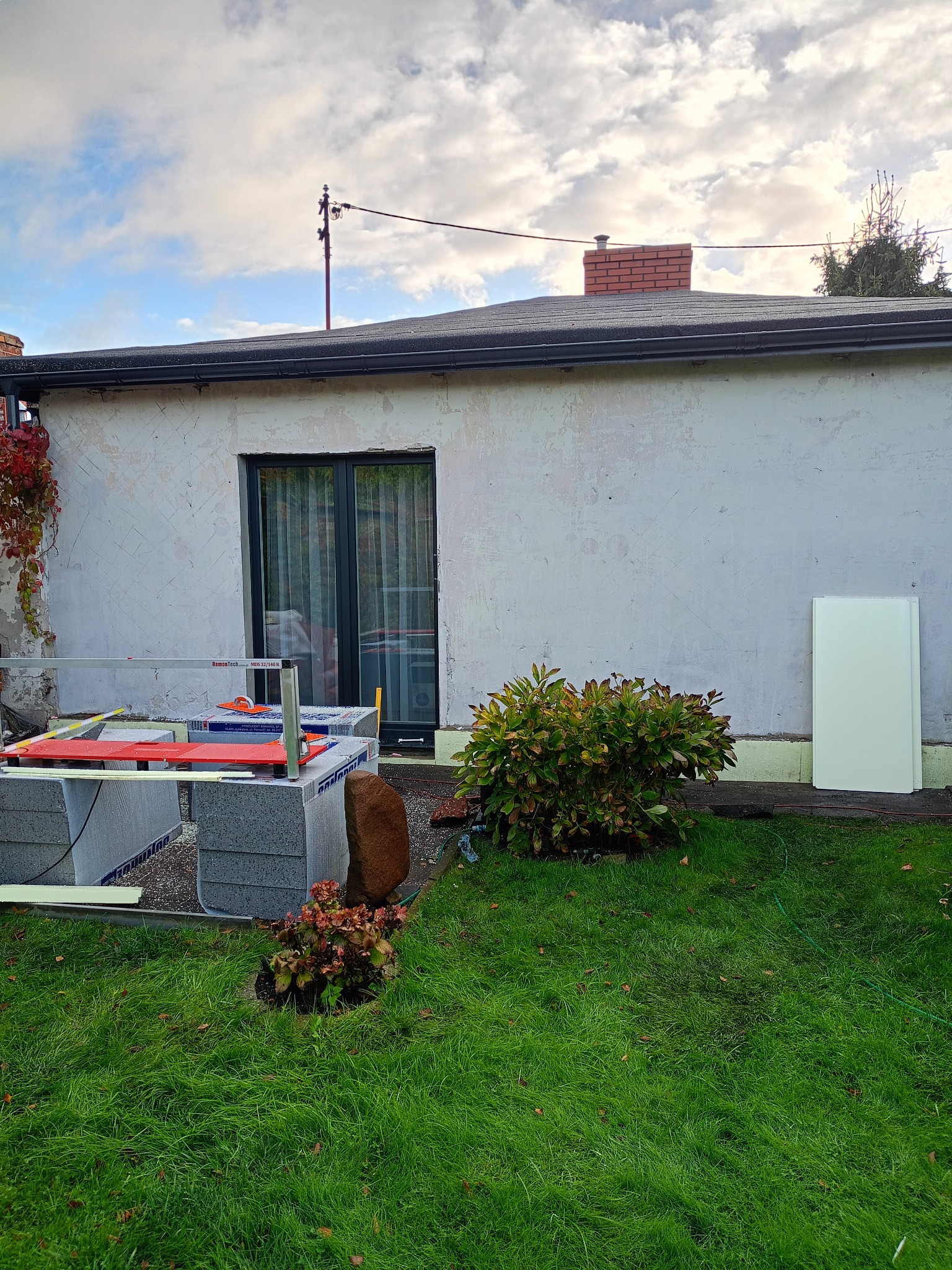 Przygotowania do ocieplenia elewacji domu jednorodzinnego. Widoczne bloczki, narzędzia i płyty izolacyjne na tle ściany z oknem i zieleni ogrodu.