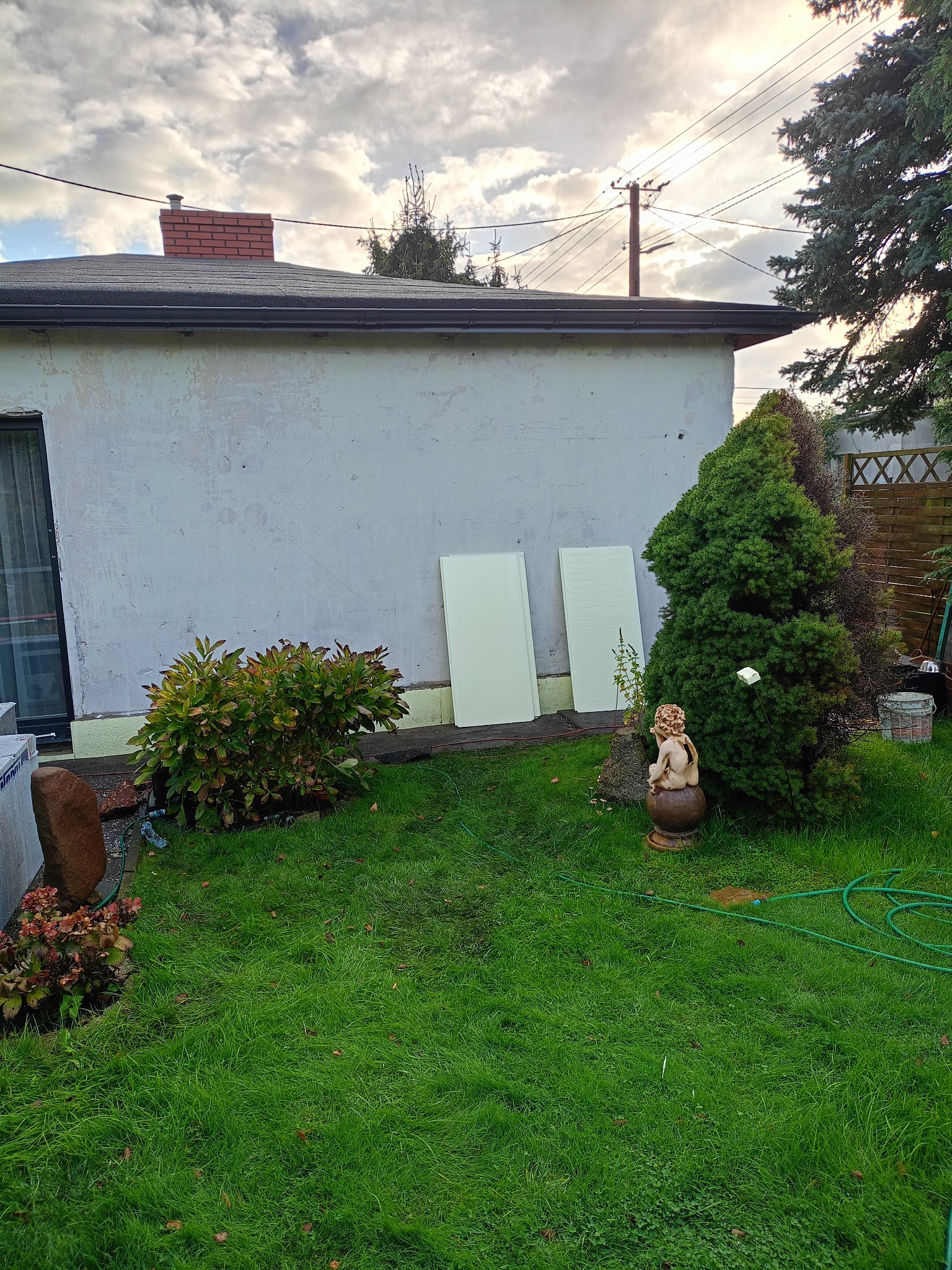 Dwie białe płyty styropianowe o fakturze, oparte o ścianę budynku z widocznymi śladami napraw, na tle zielonego trawnika i dekoracyjnego krzewu.