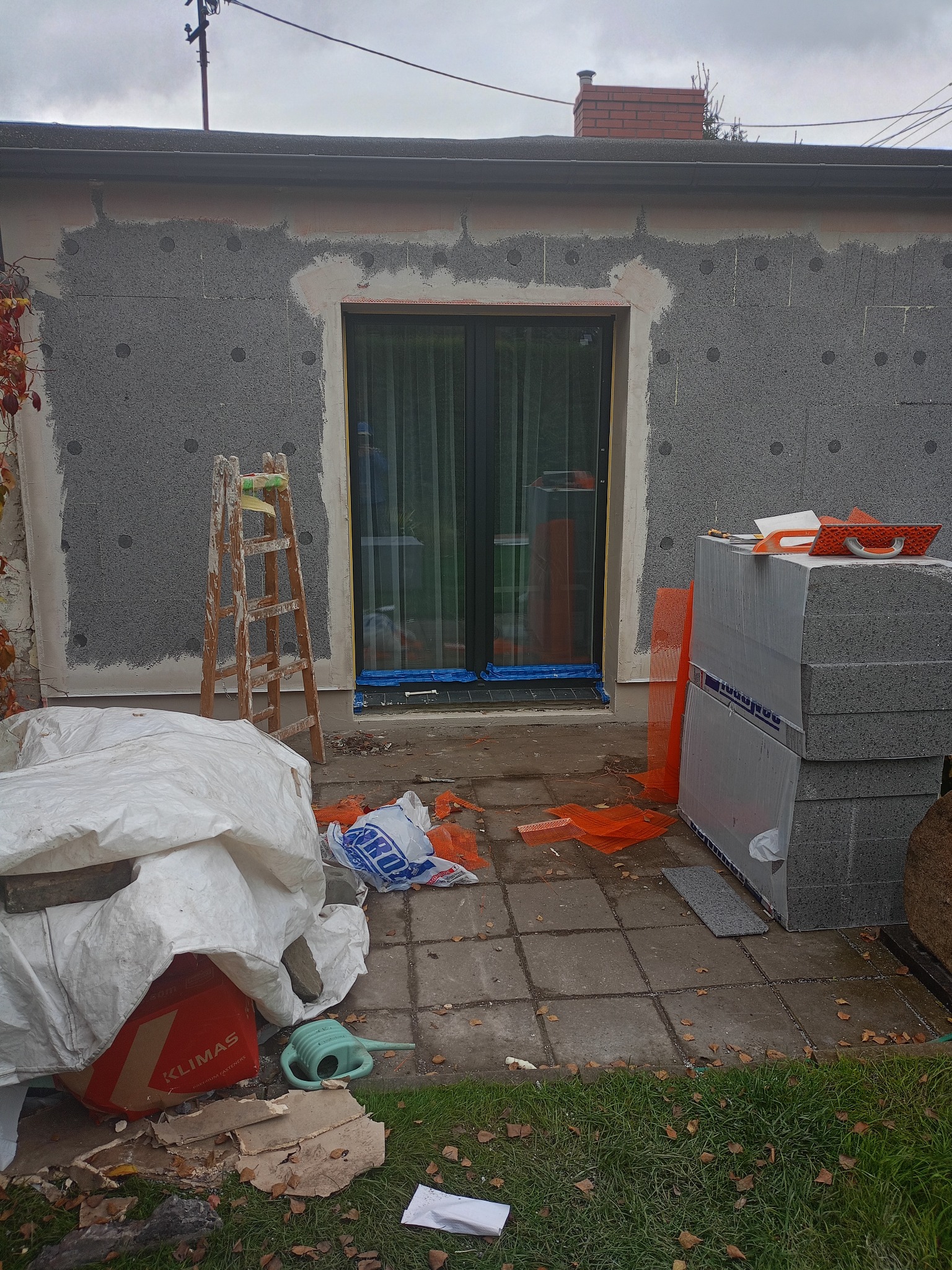 Ocieplanie ściany zewnętrznej budynku styropianem grafitowym. Widoczne rusztowanie, materiały budowlane i narzędzia. Etap prac przed nałożeniem tynku.