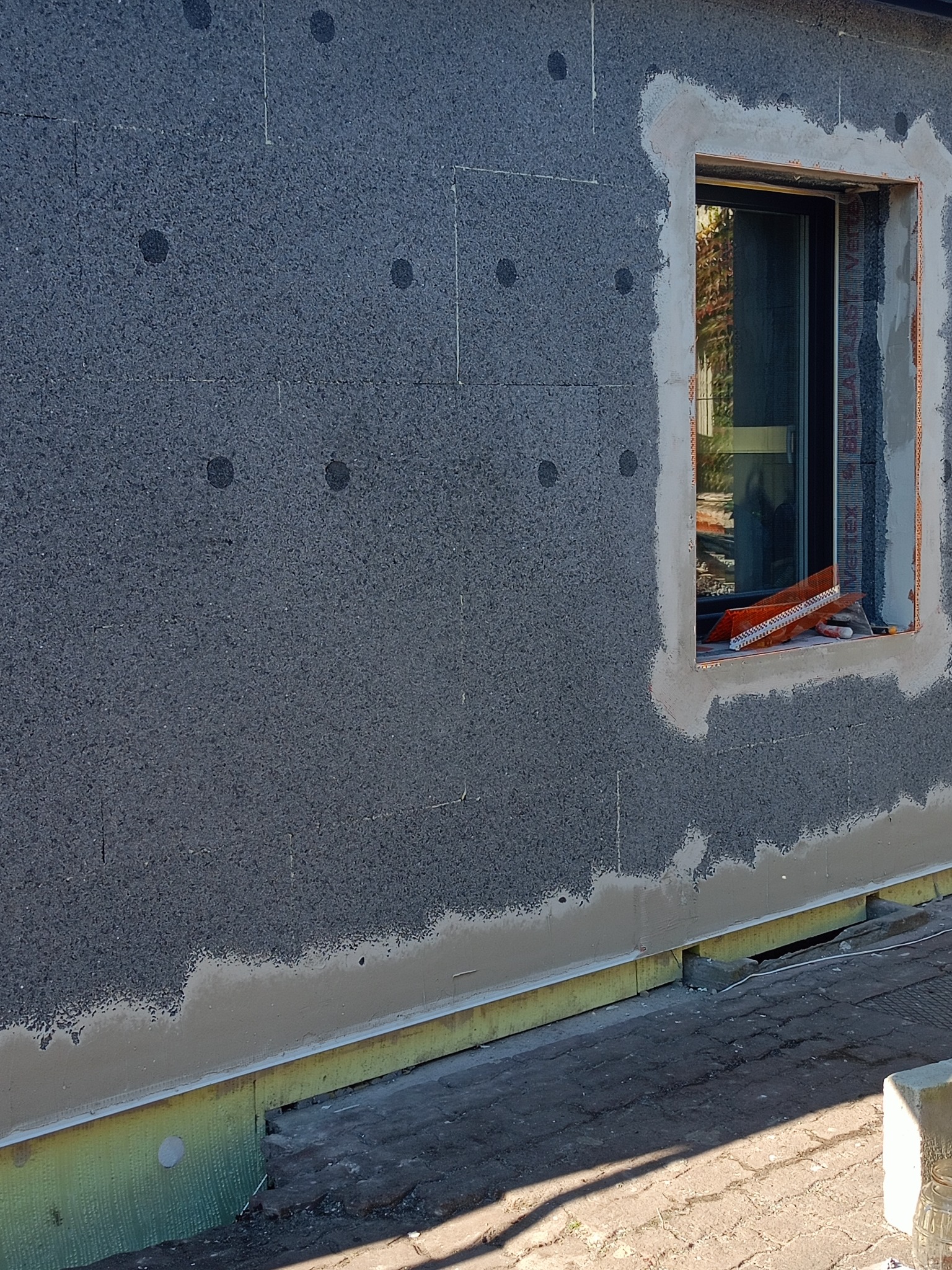 Szara elewacja budynku w trakcie ocieplania, widoczne płyty izolacyjne z czarnymi punktami, obramowanie okna w trakcie wykańczania, narzędzia w oknie.