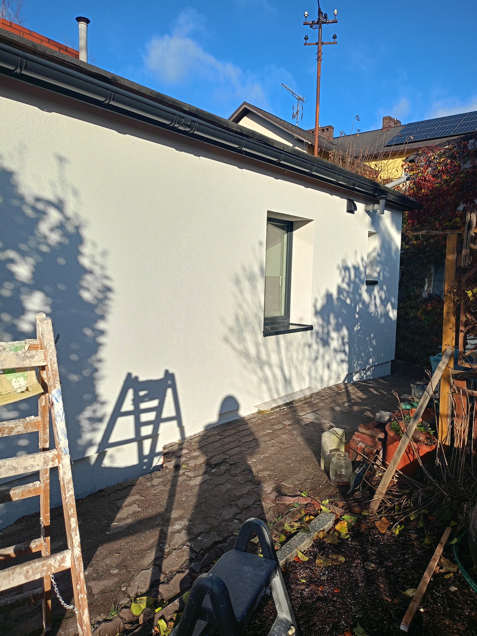 Biała elewacja budynku z czarną rynną i oknem, widoczna drabina i cień drzewa. Słoneczny dzień, błękitne niebo. Detal wykończenia.