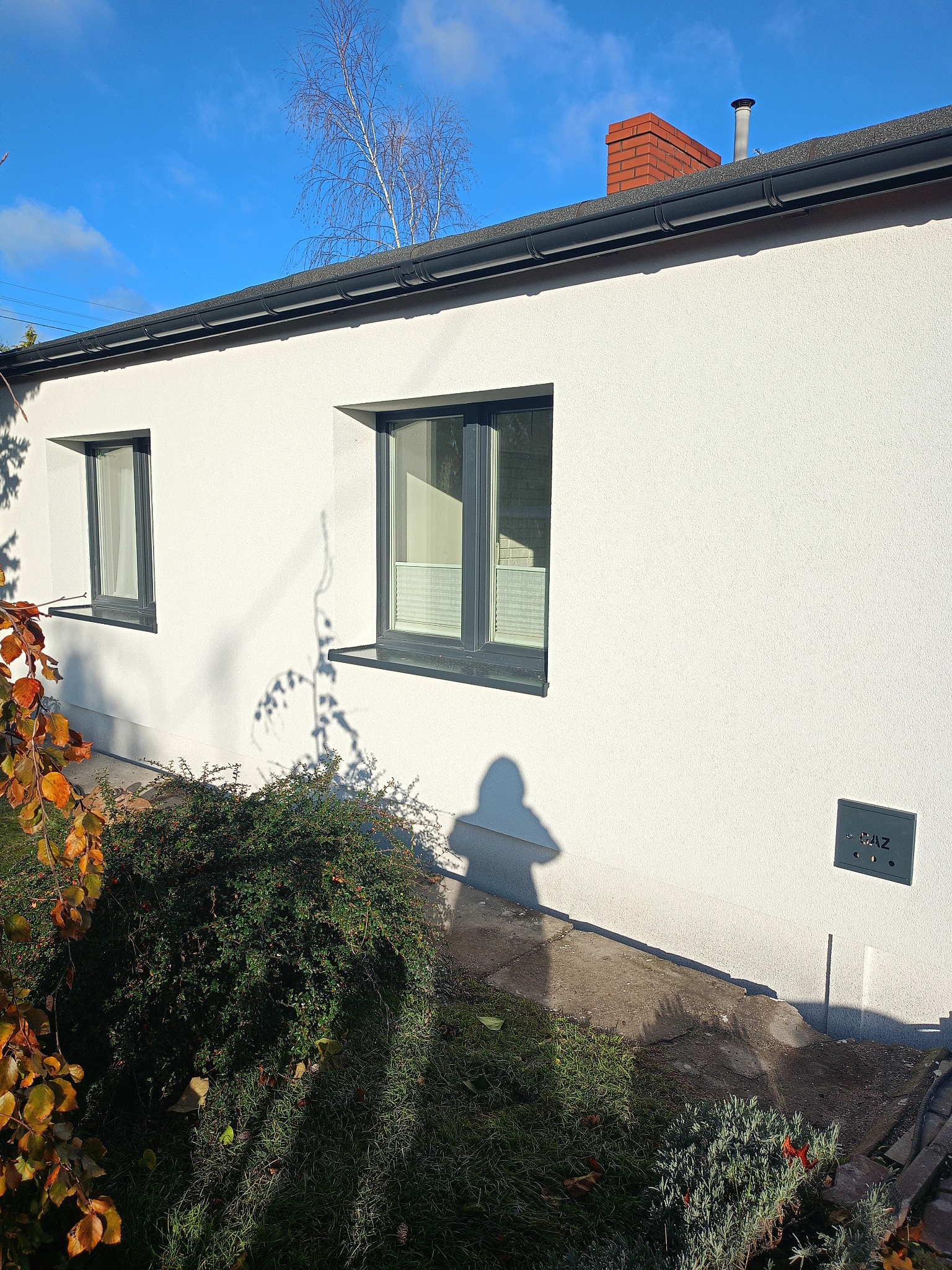 Biała elewacja domu z dwoma oknami w grafitowych ramach, widoczny fragment dachu i komin. Na pierwszym planie zieleń i cień osoby robiącej zdjęcie.