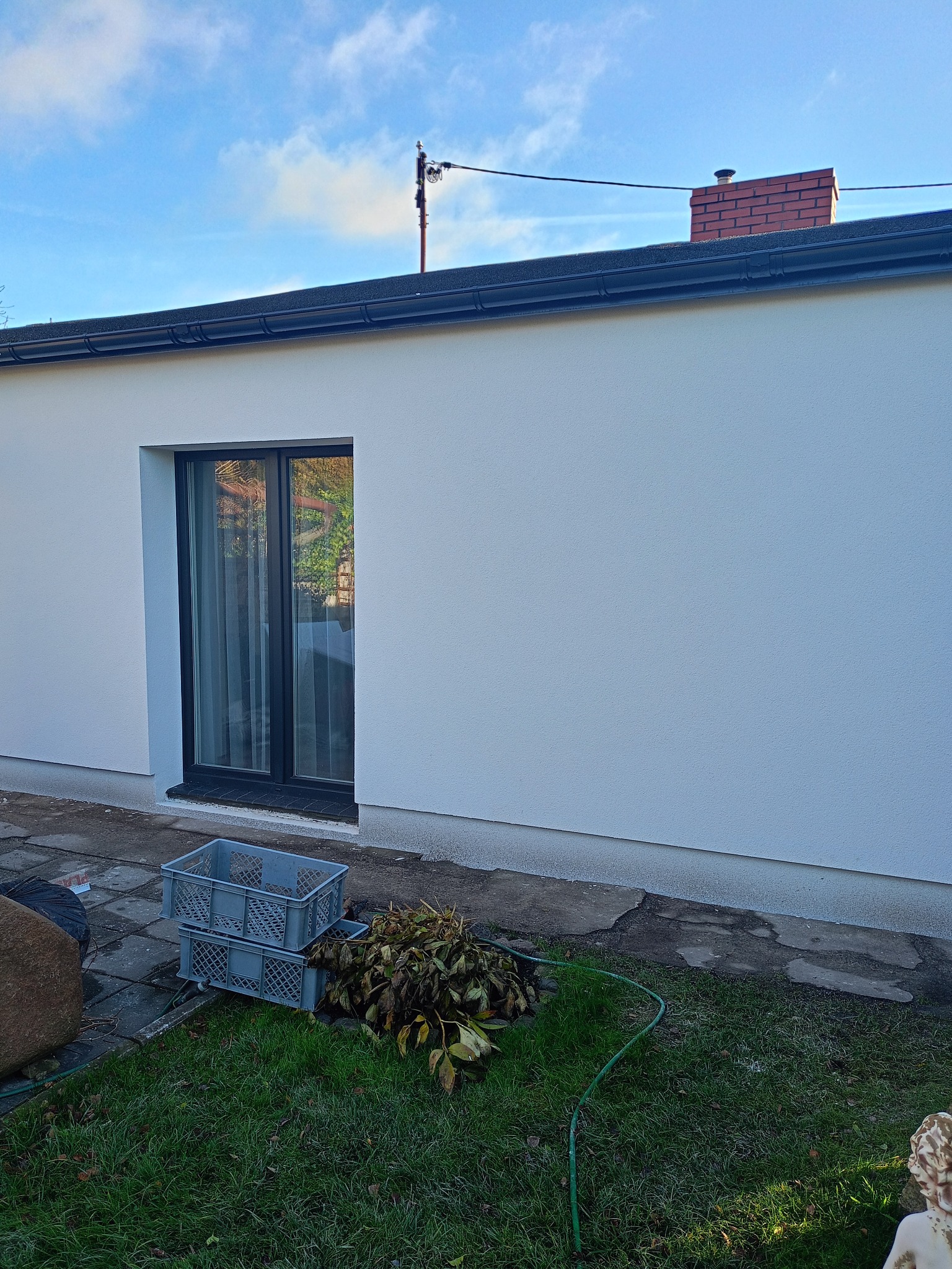 Biała elewacja domu jednorodzinnego z czarną stolarką okienną, widok z poziomu trawnika. U dołu skrzynki i zwiędłe liście.