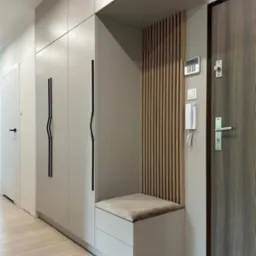 Jasna szafa wnękowa z siedziskiem i drewnianą ścianką działową przy drzwiach wejściowych, minimalistyczne czarne uchwyty.