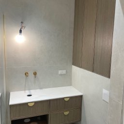 ERNEST STANIK - Nowoczesna łazienka z drewnianą szafką pod umywalkę z białym blatem i złotymi uchwytami, oraz wiszącą szafką. Szare płytki na ścianach i podwieszana toaleta.