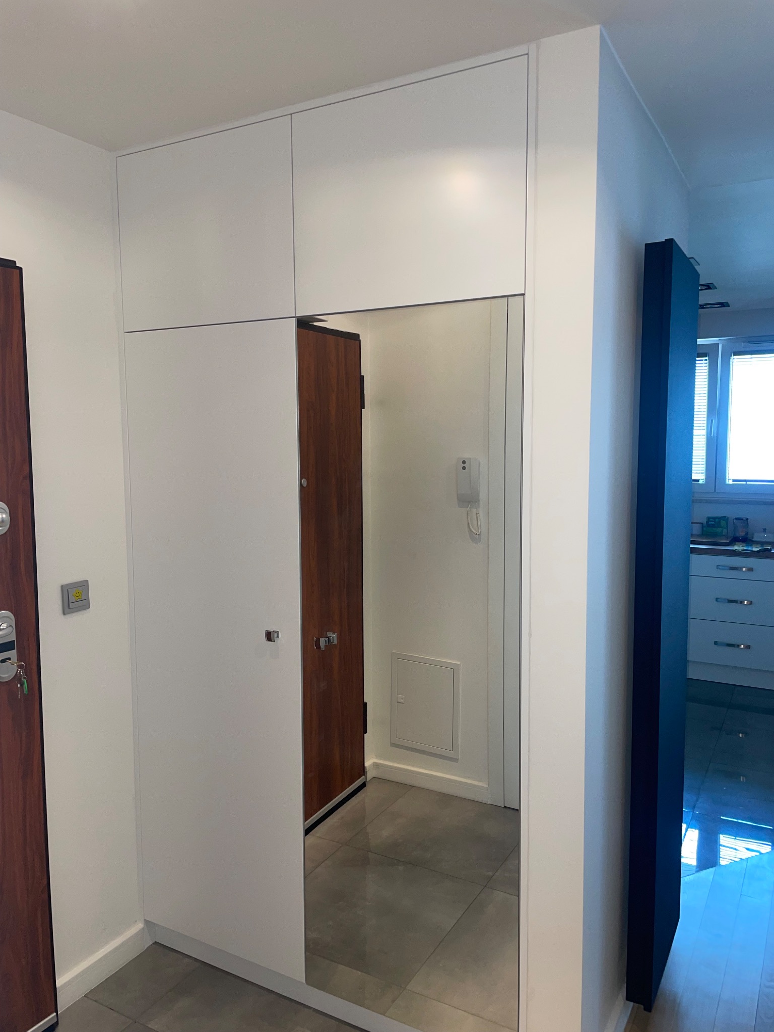 Biała zabudowa w przedpokoju z lustrzanymi drzwiami, odbijającymi wejście do innego pomieszczenia; widoczny fragment drzwi wejściowych po lewej stronie.