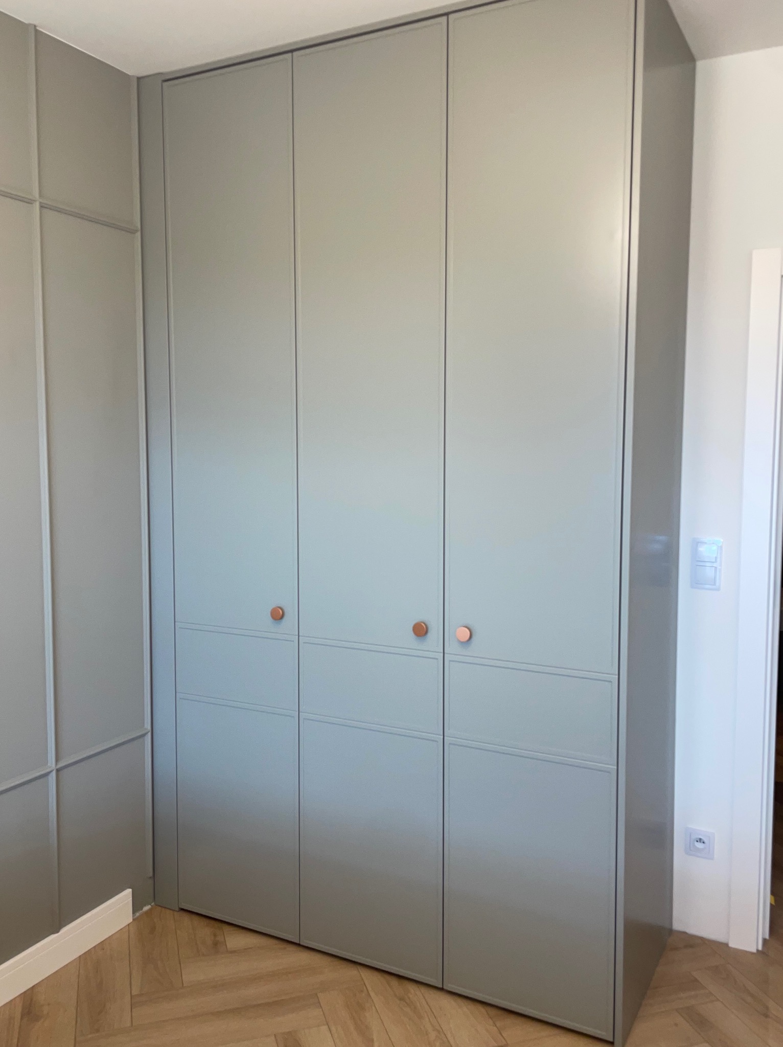 Jasnoszara szafa wnękowa z trzema parami drzwi, z miedzianymi gałkami, dopasowana do ściany z dekoracyjnymi listwami i parkietem w jodełkę.