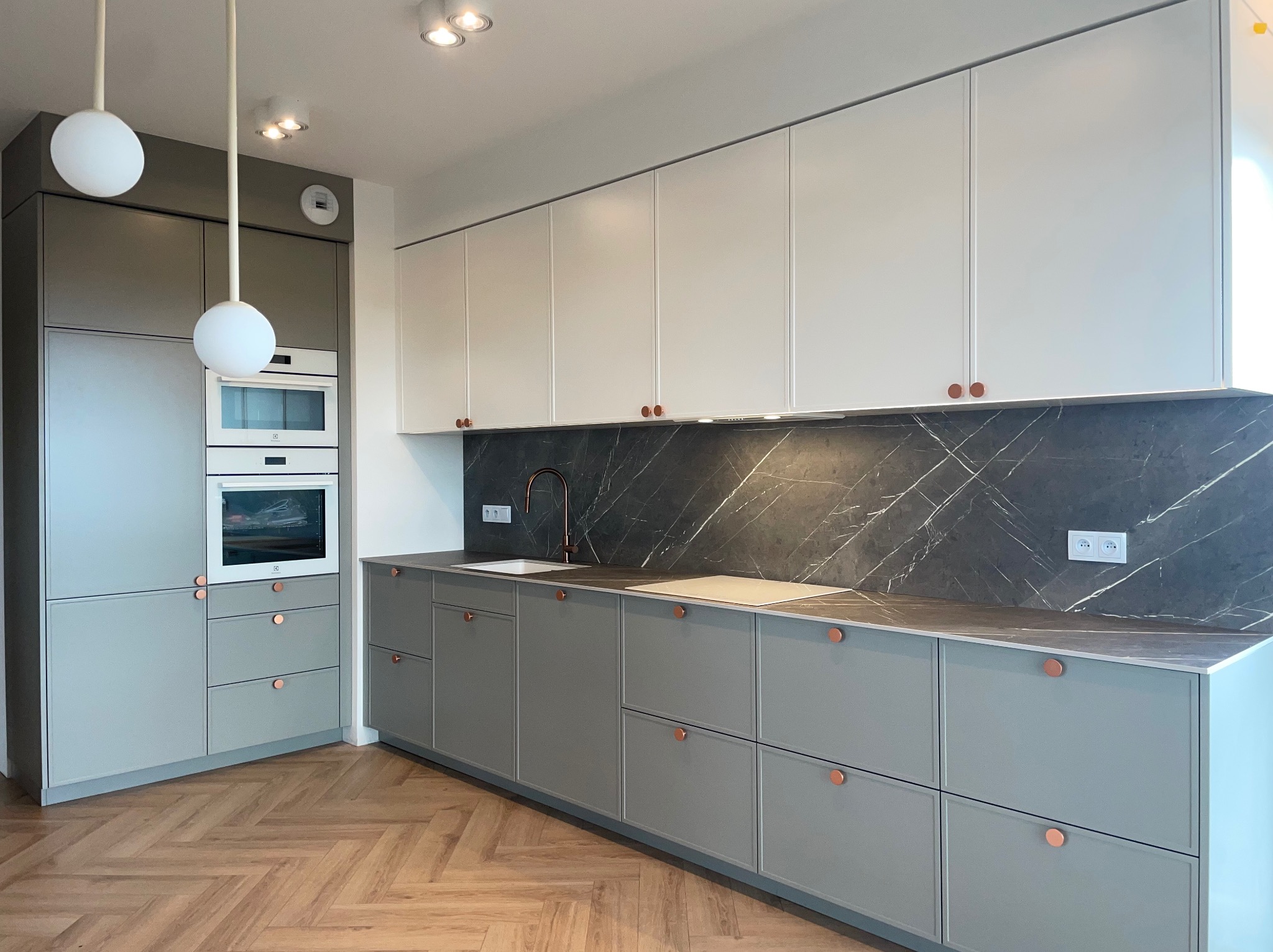 Nowoczesna kuchnia z szarymi dolnymi szafkami i białymi górnymi, marmurowy backsplash, drewniana podłoga w jodełkę i okrągłe lampy wiszące.