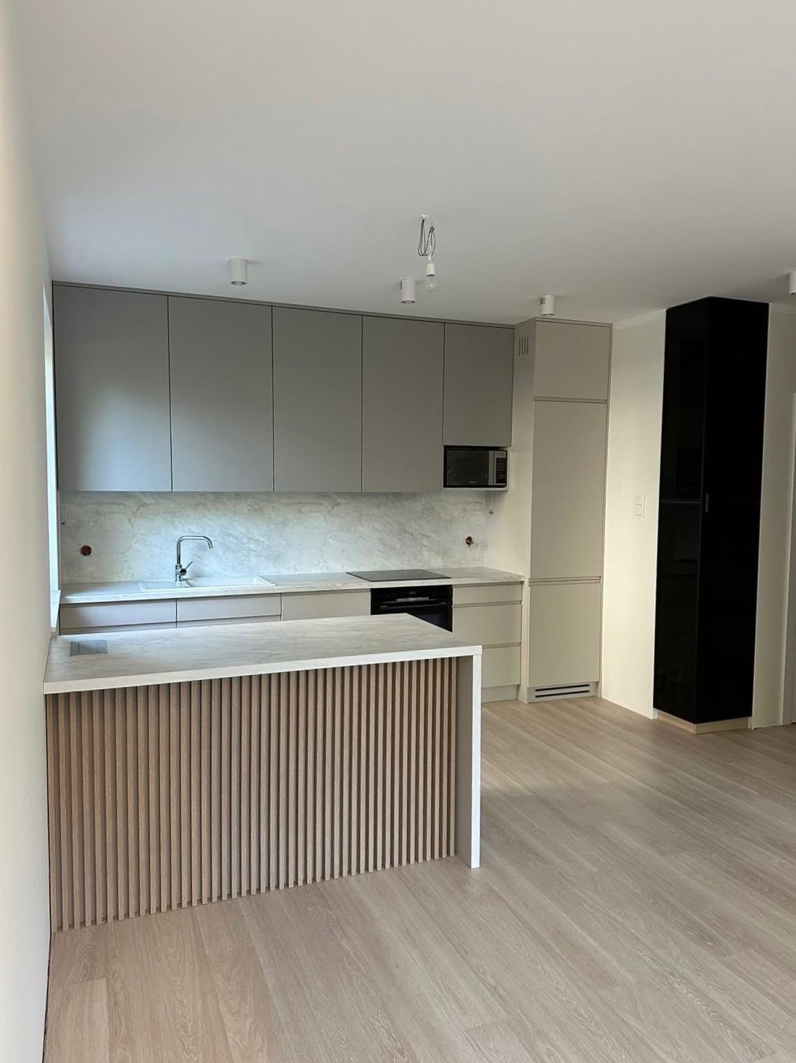 Jasna kuchnia z wyspą wykończoną drewnianymi lamelami, szare szafki, marmurowy backsplash i czarny słupek. Minimalistyczny design, nowoczesne oświetlenie.