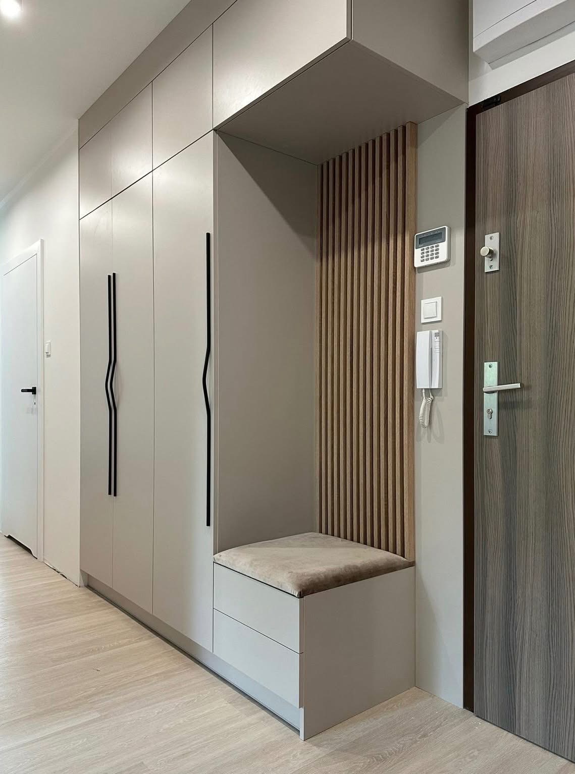 Jasna szafa wnękowa z siedziskiem i drewnianą ścianką działową przy drzwiach wejściowych, minimalistyczne czarne uchwyty.