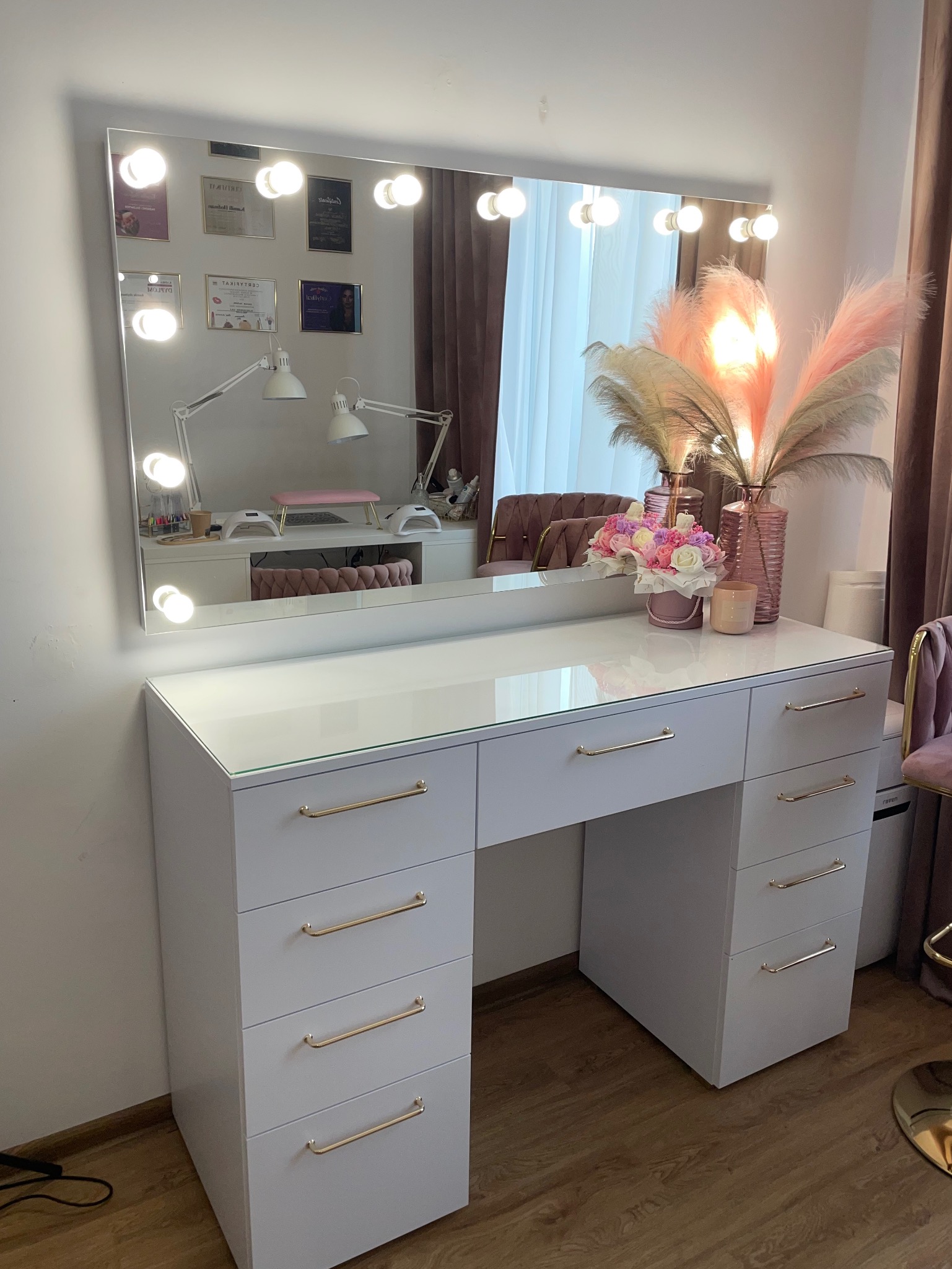 Biała toaletka z lustrem podświetlanym żarówkami, złote uchwyty szuflad, kwiaty i trawa pampasowa jako dekoracja. W lustrze odbicie stanowiska do manicure.