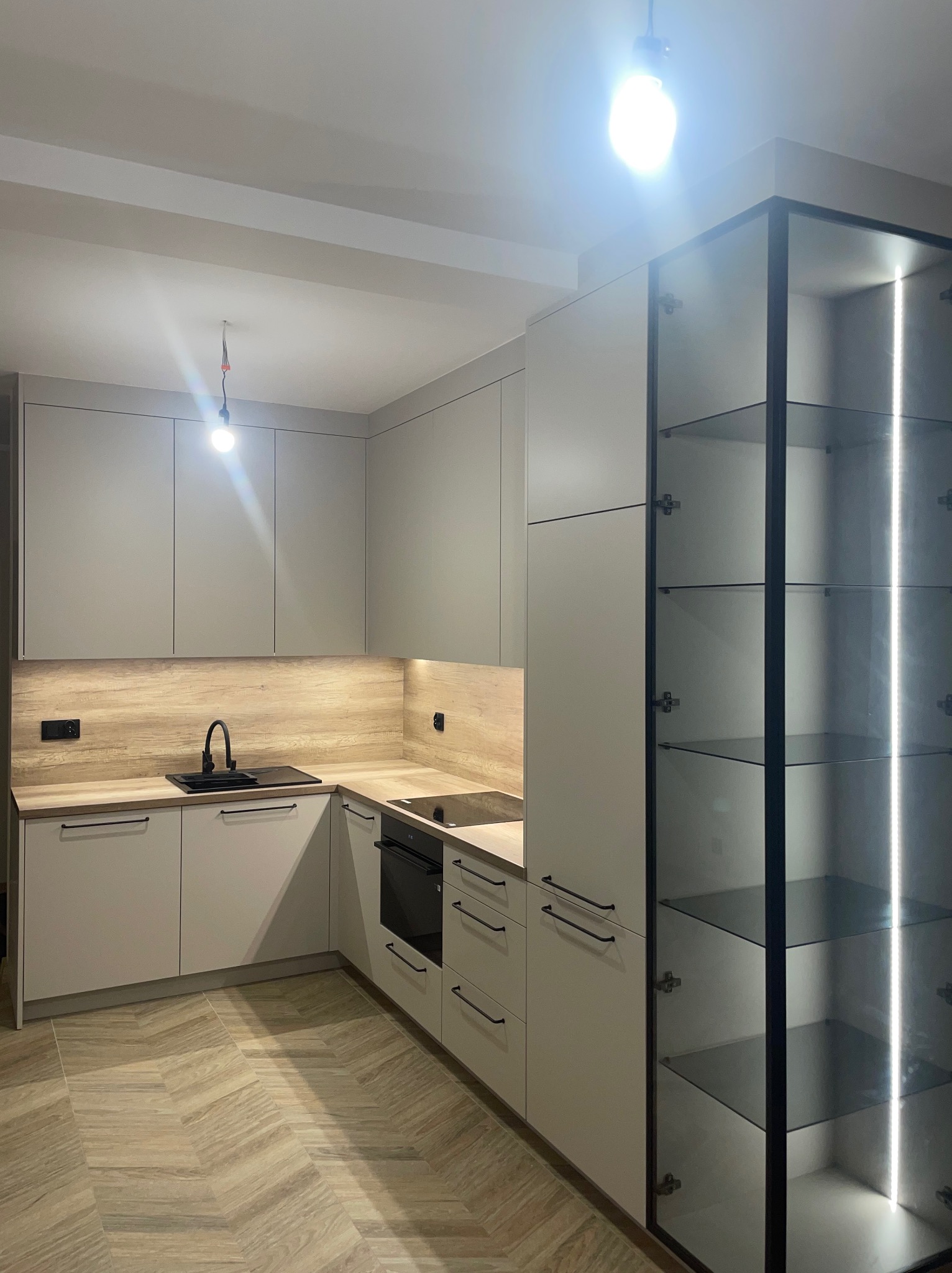 Jasna kuchnia z drewnianym blatem i czarną armaturą. Szklana witryna z oświetleniem LED. Parkiet ułożony w jodełkę. Nowoczesny design.