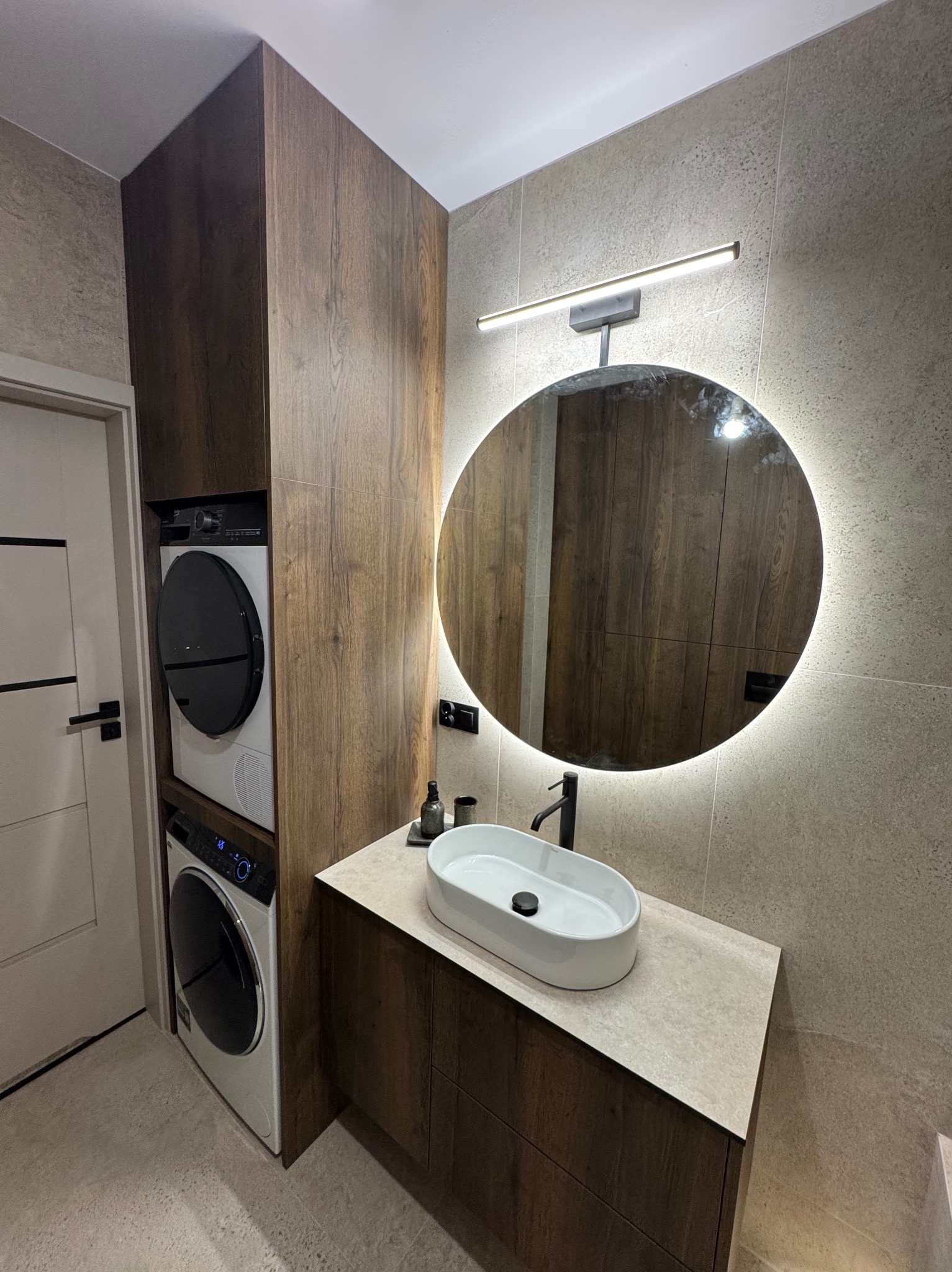 Nowoczesna łazienka z drewnianą zabudową, okrągłym lustrem z podświetleniem LED, umywalką nablatową i pralko-suszarką wkomponowaną w szafę.
