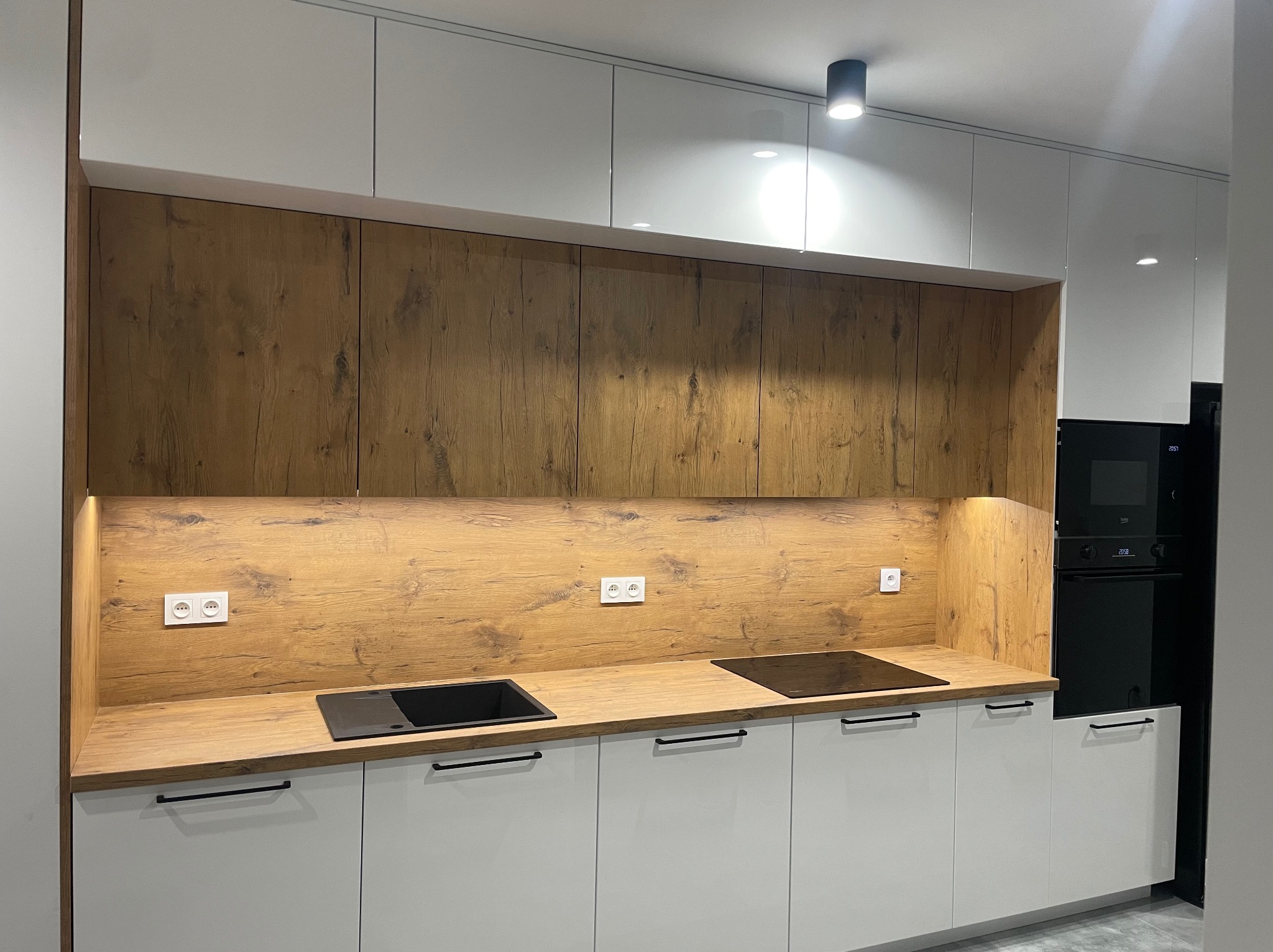 Nowoczesna kuchnia z białymi szafkami dolnymi i górnymi, drewnianym blatem i panelem ściennym. Czarny piekarnik w zabudowie. Oświetlenie LED pod szafkami górnymi.