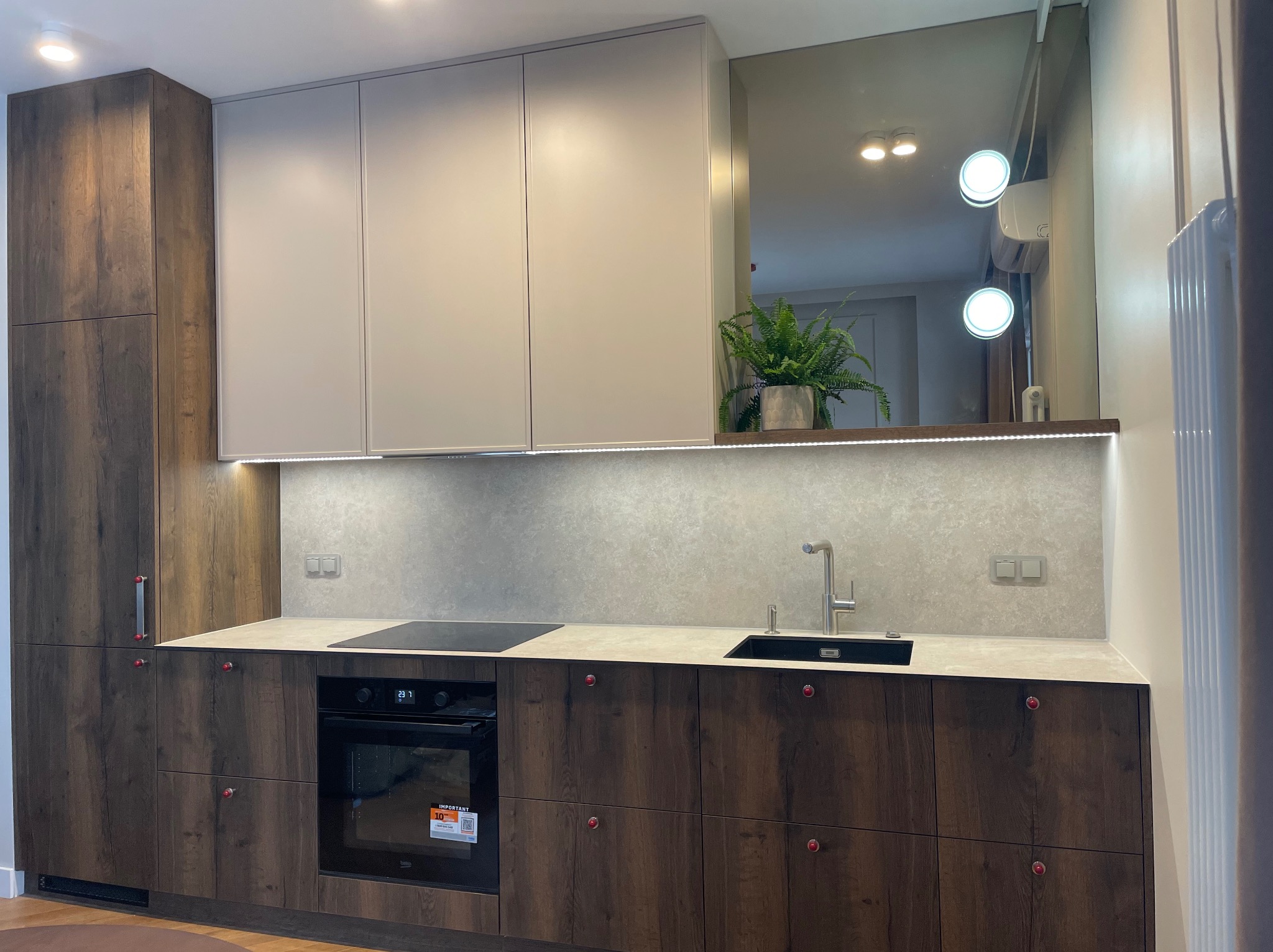 Nowoczesna kuchnia z drewnianymi szafkami, beżowymi górnymi szafkami i lustrem z rośliną. Oświetlenie LED pod szafkami górnymi. Minimalistyczny design.