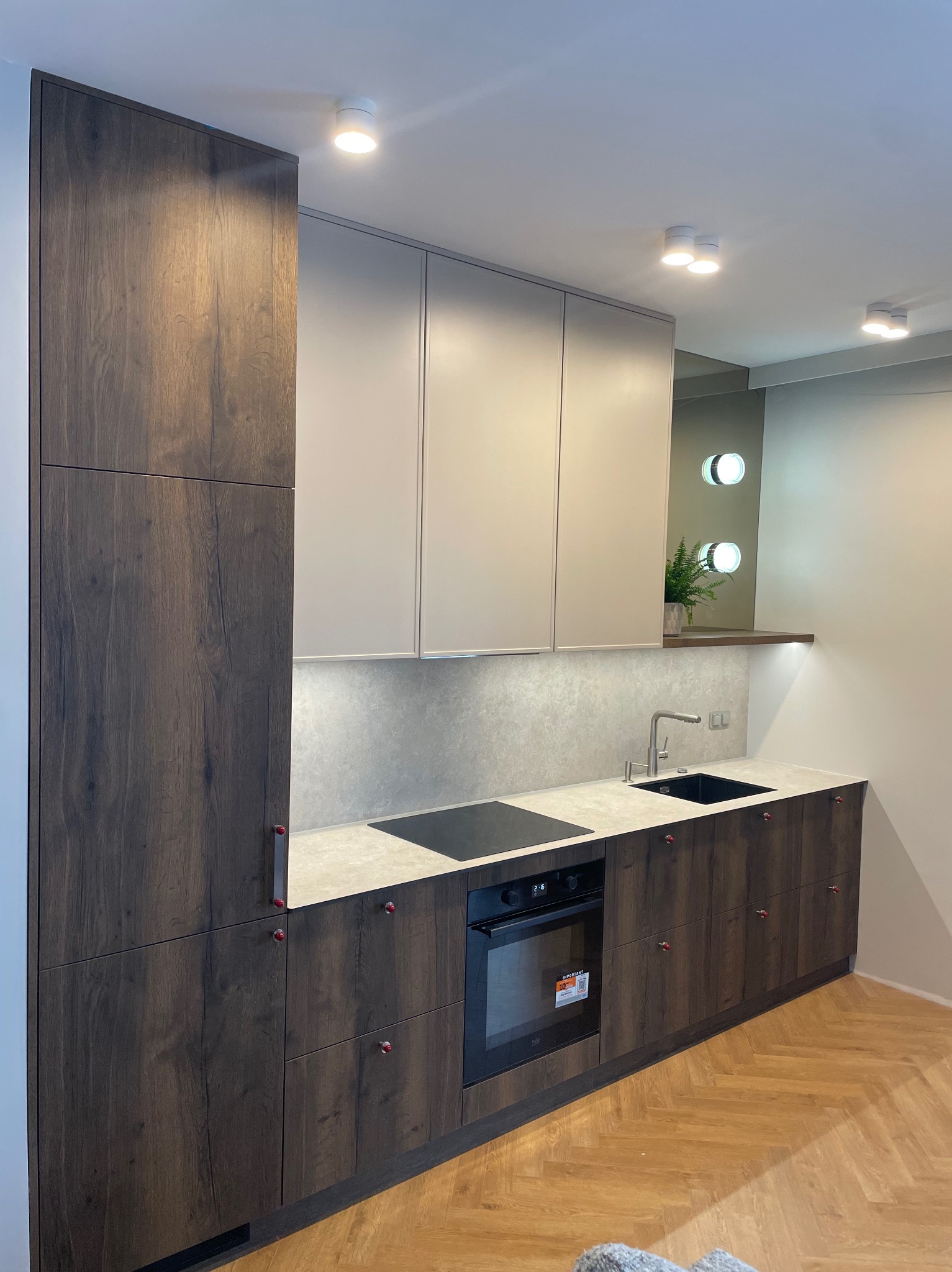 Nowoczesna kuchnia na wymiar z ciemnego drewna i jasnymi górnymi szafkami. Uwagę przykuwają czerwone gałki i parkiet ułożony w jodełkę.