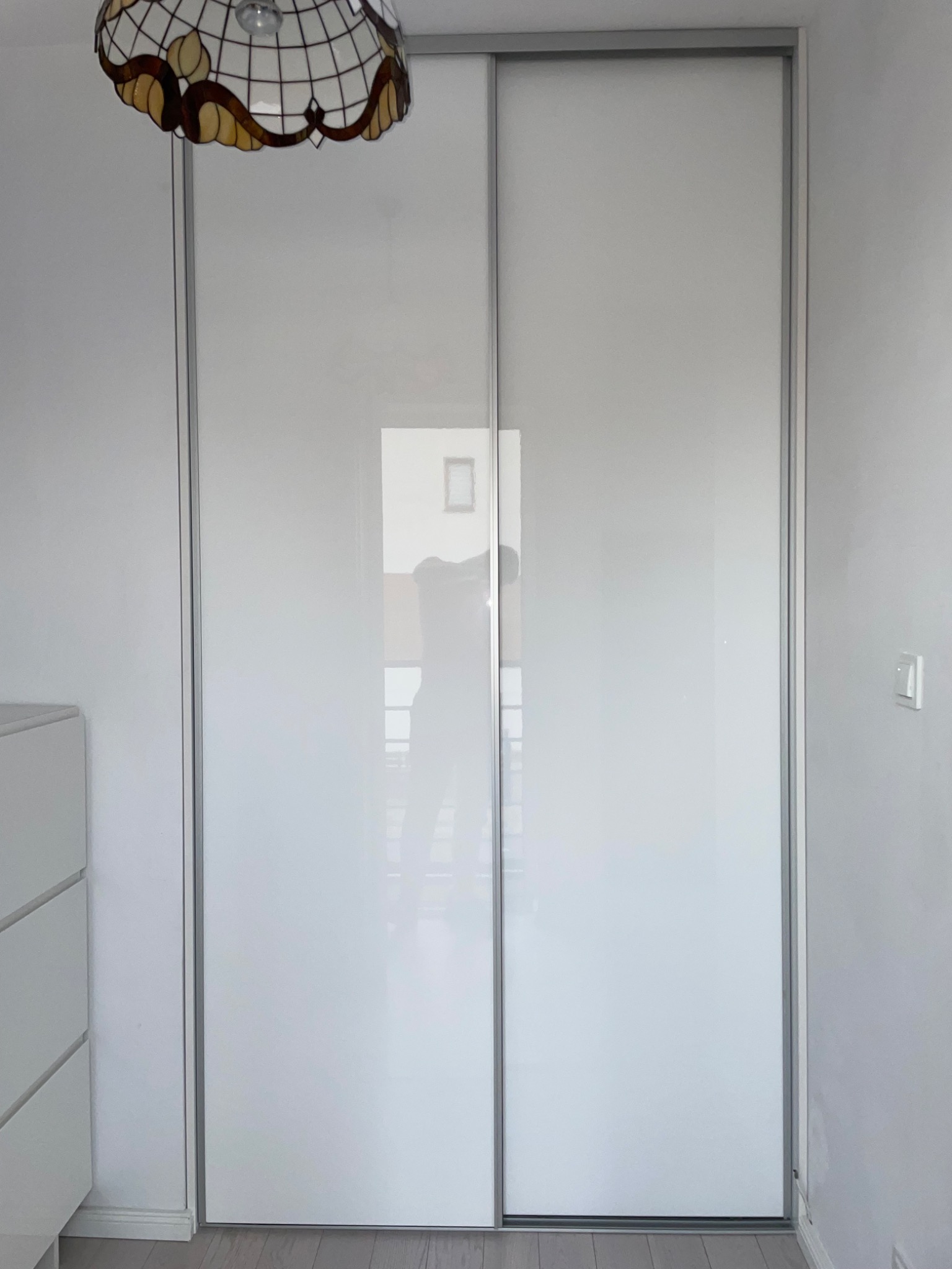 Biała szafa przesuwna z lustrzanymi drzwiami, widoczna w odbiciu sylwetka osoby, nad szafą lampa z witrażowym kloszem, obok komoda z szufladami.