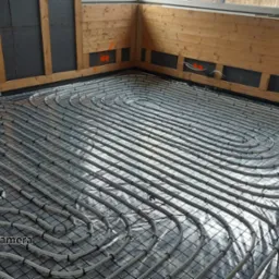 Białe rury ogrzewania podłogowego ułożone spiralnie na czarnej siatce montażowej, widoczne izolowane ściany i drewniane wykończenie w nowym budynku.