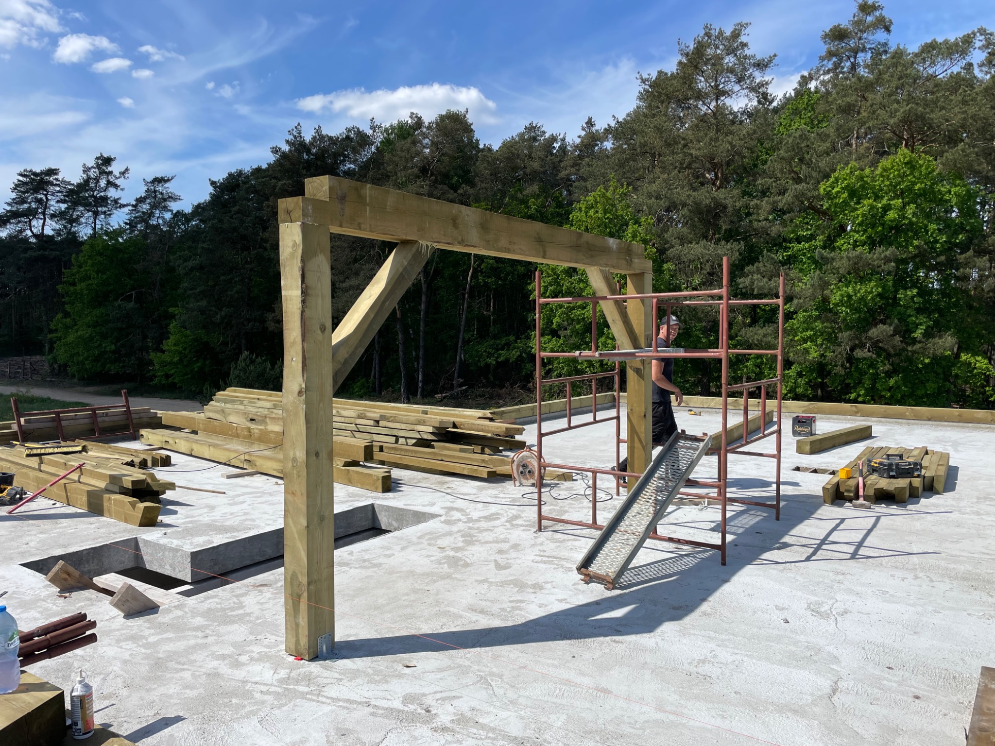 Wznoszenie drewnianej konstrukcji szkieletowej na betonowej płycie fundamentowej, widoczne rusztowanie i przygotowane elementy drewniane, w tle las.