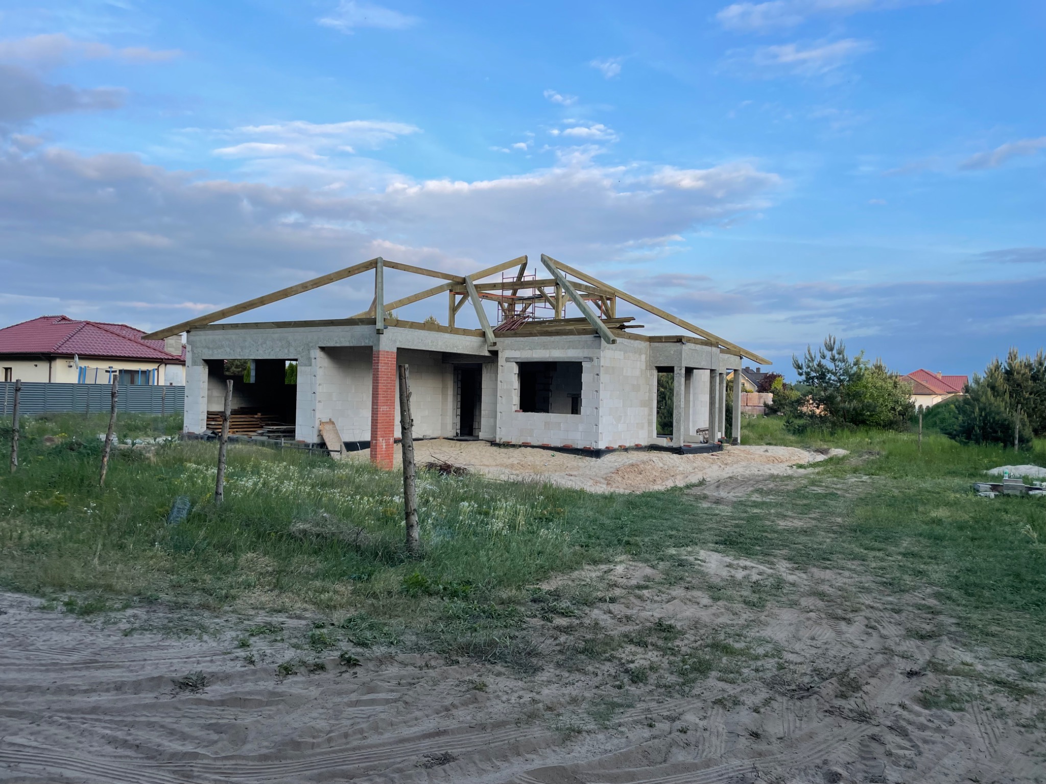 Niedokończony budynek jednorodzinny z betonu komórkowego z częściowo zmontowaną drewnianą konstrukcją dachu, widoczny z zewnątrz na tle nieba z chmurami.
