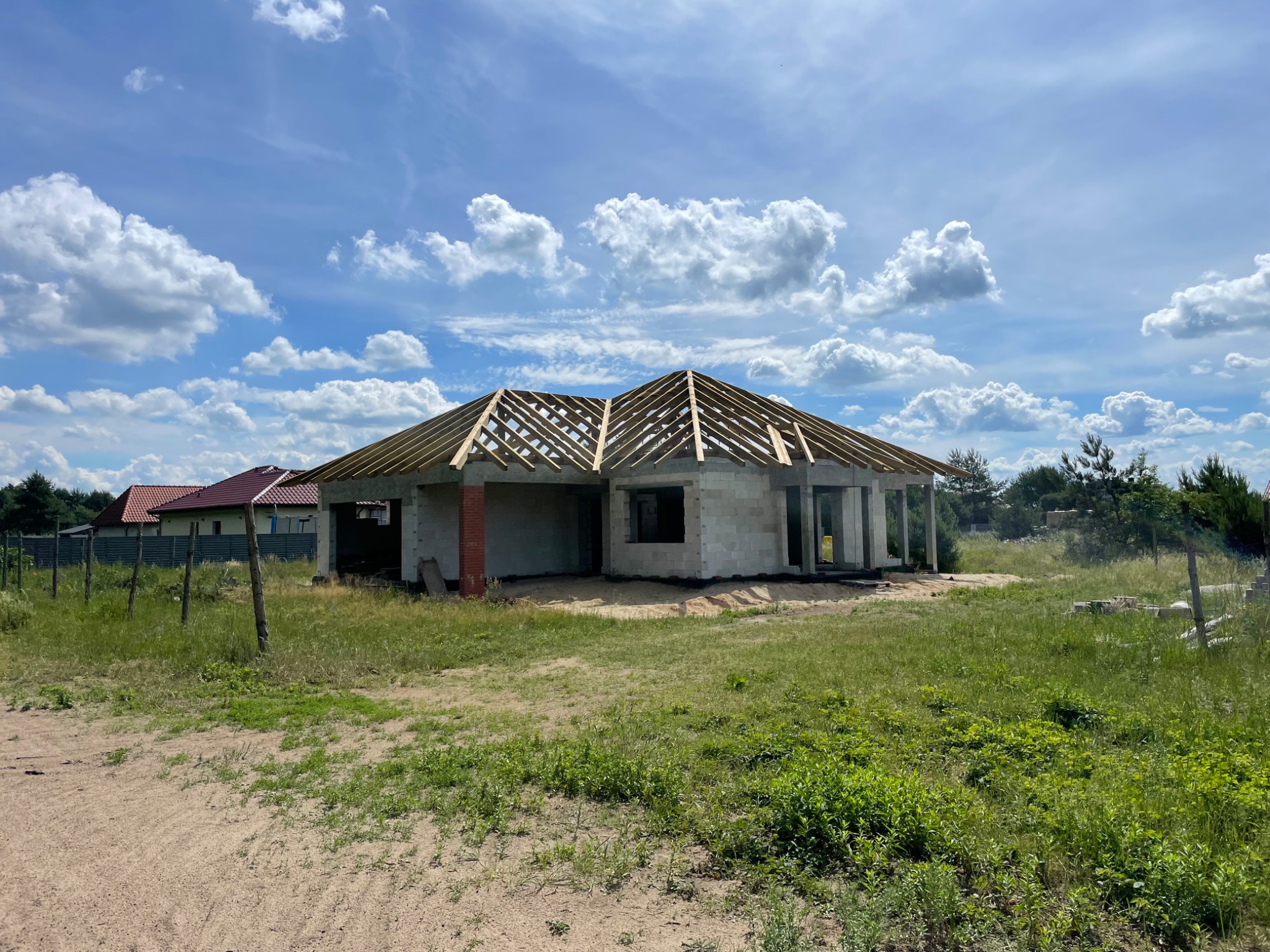 Surowy stan domu murowanego z białych bloczków, widoczne drewniane elementy konstrukcji dachu na tle błękitnego nieba z obłokami.