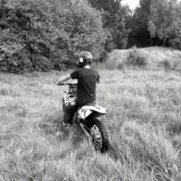 Czarno-białe zdjęcie motocykla crossowego z jeźdźcem w kasku na zarośniętym terenie, widok z tyłu, wysoka trawa i drzewa w tle.