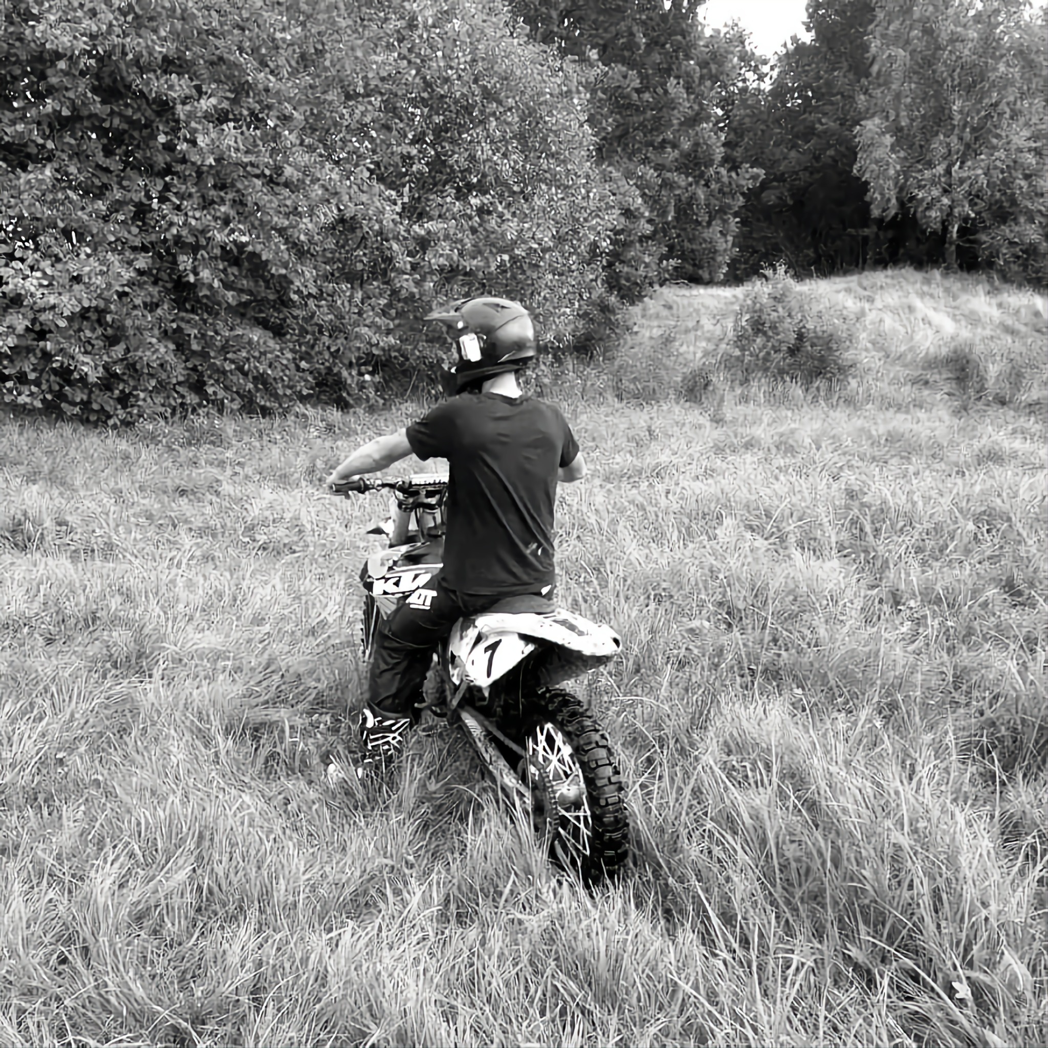Czarno-białe zdjęcie motocykla crossowego z jeźdźcem w kasku na zarośniętym terenie, widok z tyłu, wysoka trawa i drzewa w tle.