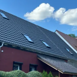 Ciemnoszary dach z dachówki ceramicznej z oknami połaciowymi, widok z boku domu w kolorze bordowym, z widocznymi rynnami i fragmentami innych budynków w tle pod błękitnym niebem z białymi chmurami.