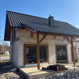 Andrzej Geburek - Dom w trakcie budowy z ciemną dachówką i drewnianą konstrukcją werandy. Na dachu widoczny komin z ozdobną figurką. Wokół budynku gruz i materiały budowlane.