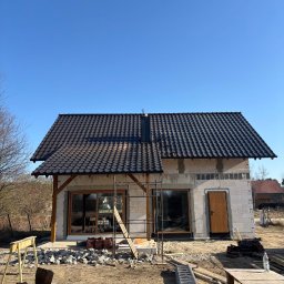 Andrzej Geburek - Dom jednorodzinny w trakcie budowy, z ciemną dachówką ceramiczną i widoczną konstrukcją więźby. Na pierwszym planie materiały budowlane i rusztowania.