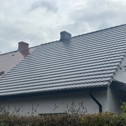 Andrzej Geburek - Nowy, ciemnoszary dach z dachówki bitumicznej na białym domu jednorodzinnym, widok z dołu, z fragmentem sąsiedniego dachu z ceglaną dachówką i kominem.
