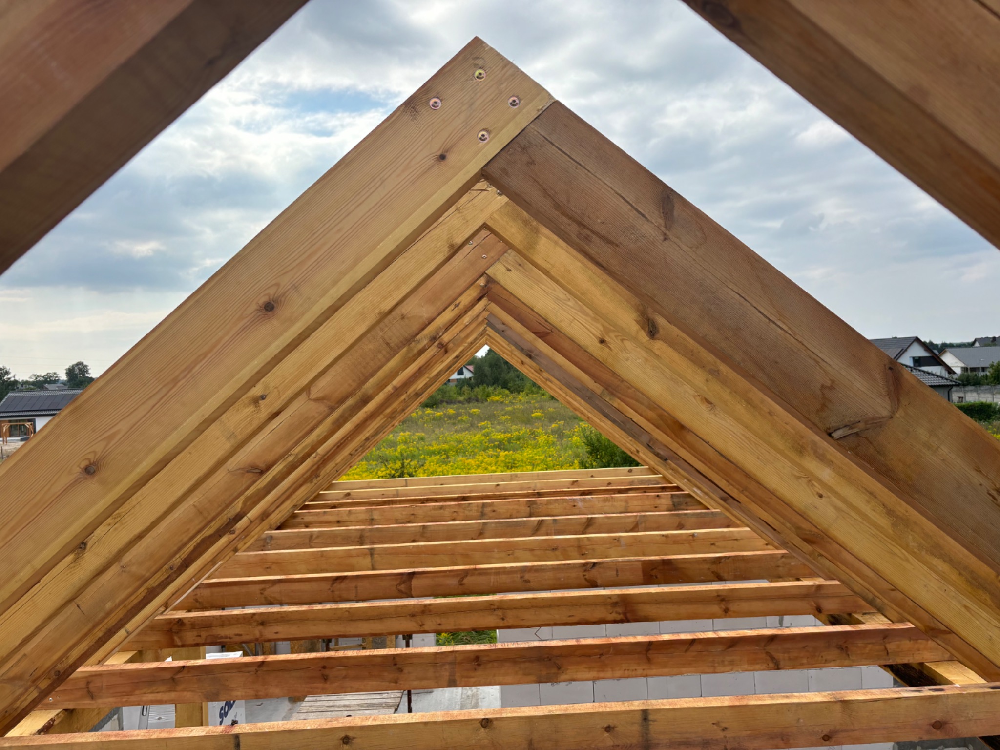 Drewniana konstrukcja więźby dachowej z widocznymi elementami łączeń na tle błękitnego nieba i zieleni w oddali.