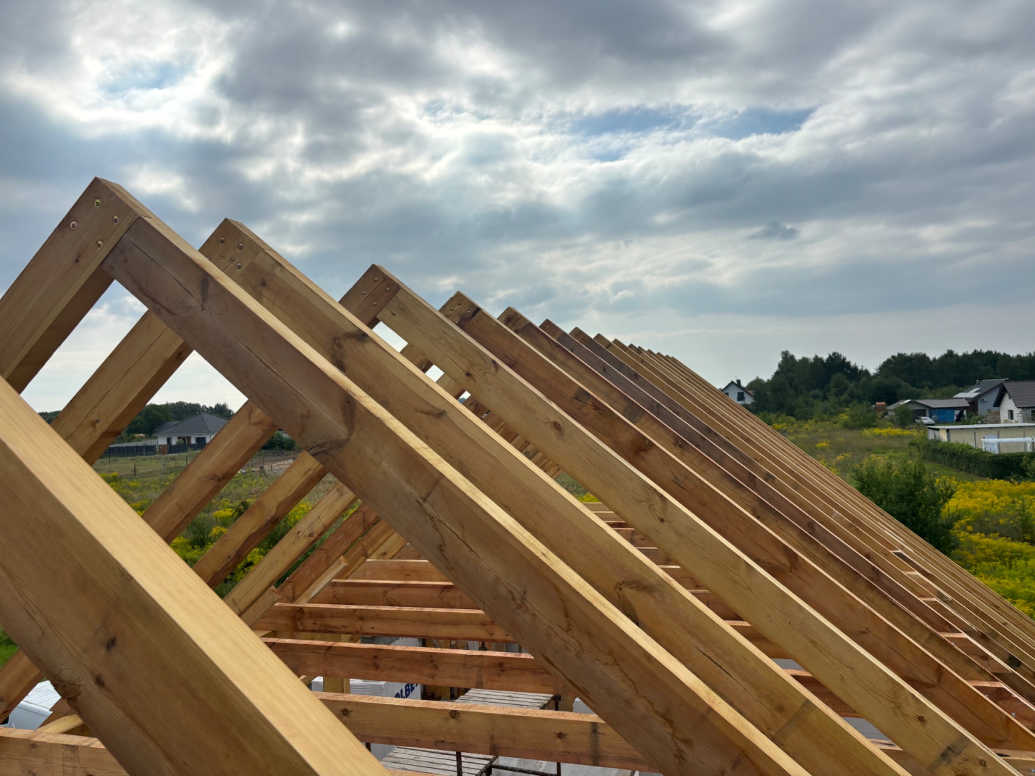 Surowa drewniana konstrukcja dachu z widocznymi elementami łączącymi, na tle zachmurzonego nieba i zabudowań wiejskich.