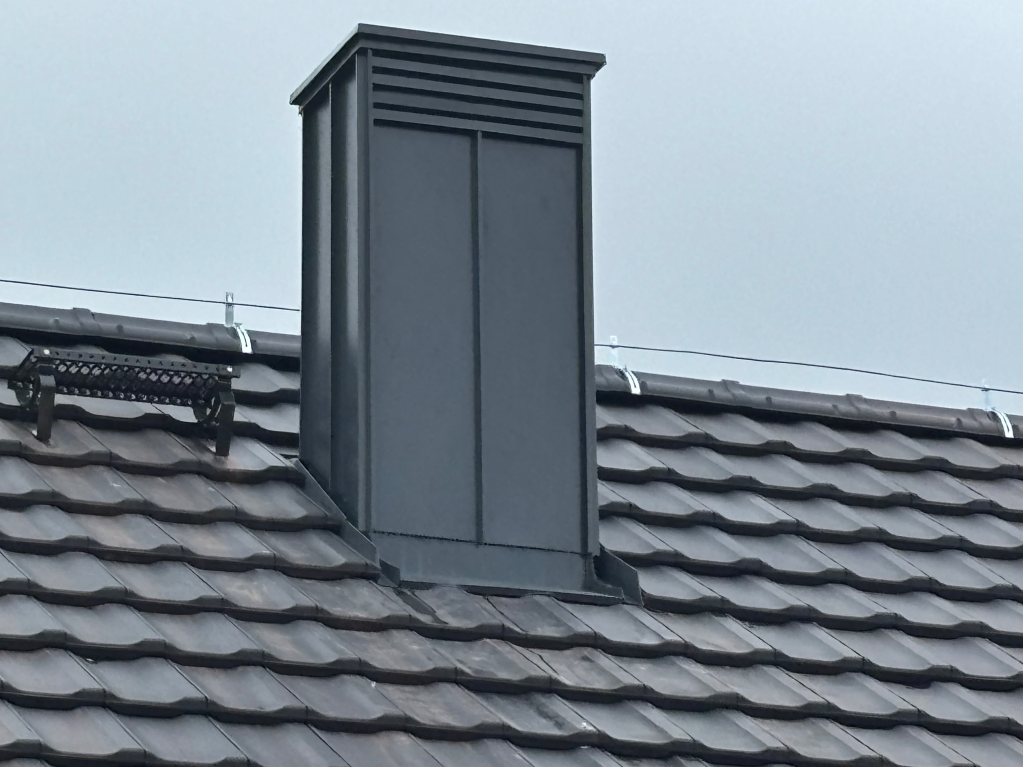 Szary komin wentylacyjny z poziomymi szczelinami na dachu pokrytym ciemną dachówką ceramiczną, widoczny uchwyt do montażu drabiny dachowej.